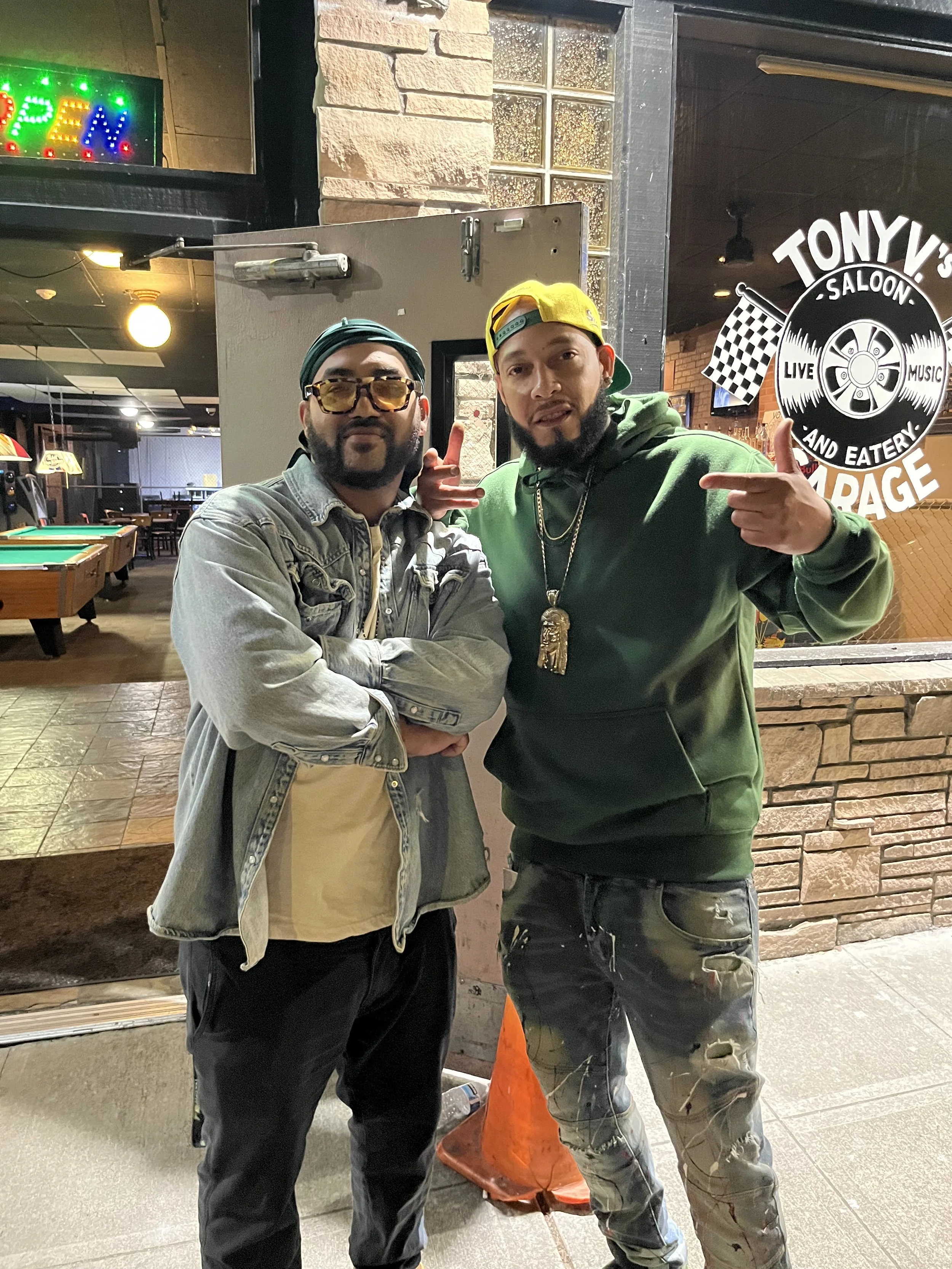 Two men standing outside Tony's Saloon, with one wearing a denim jacket, sunglasses, and a green bandana, and the other wearing a green hoodie, yellow cap, and large chain necklace, both posing confidently.