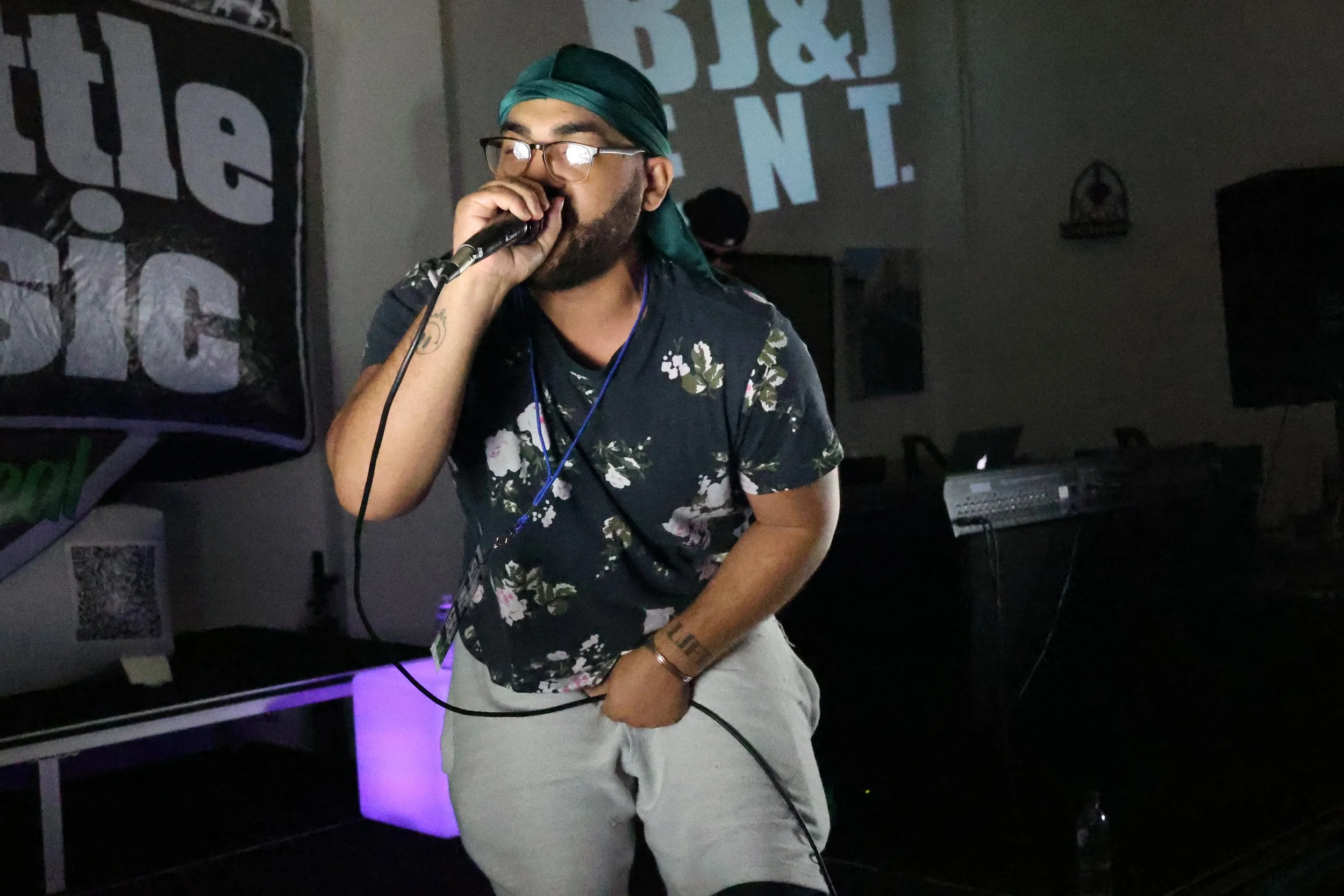 Man wearing glasses, a green durag, and a black floral shirt, singing into a microphone at an indoor event.