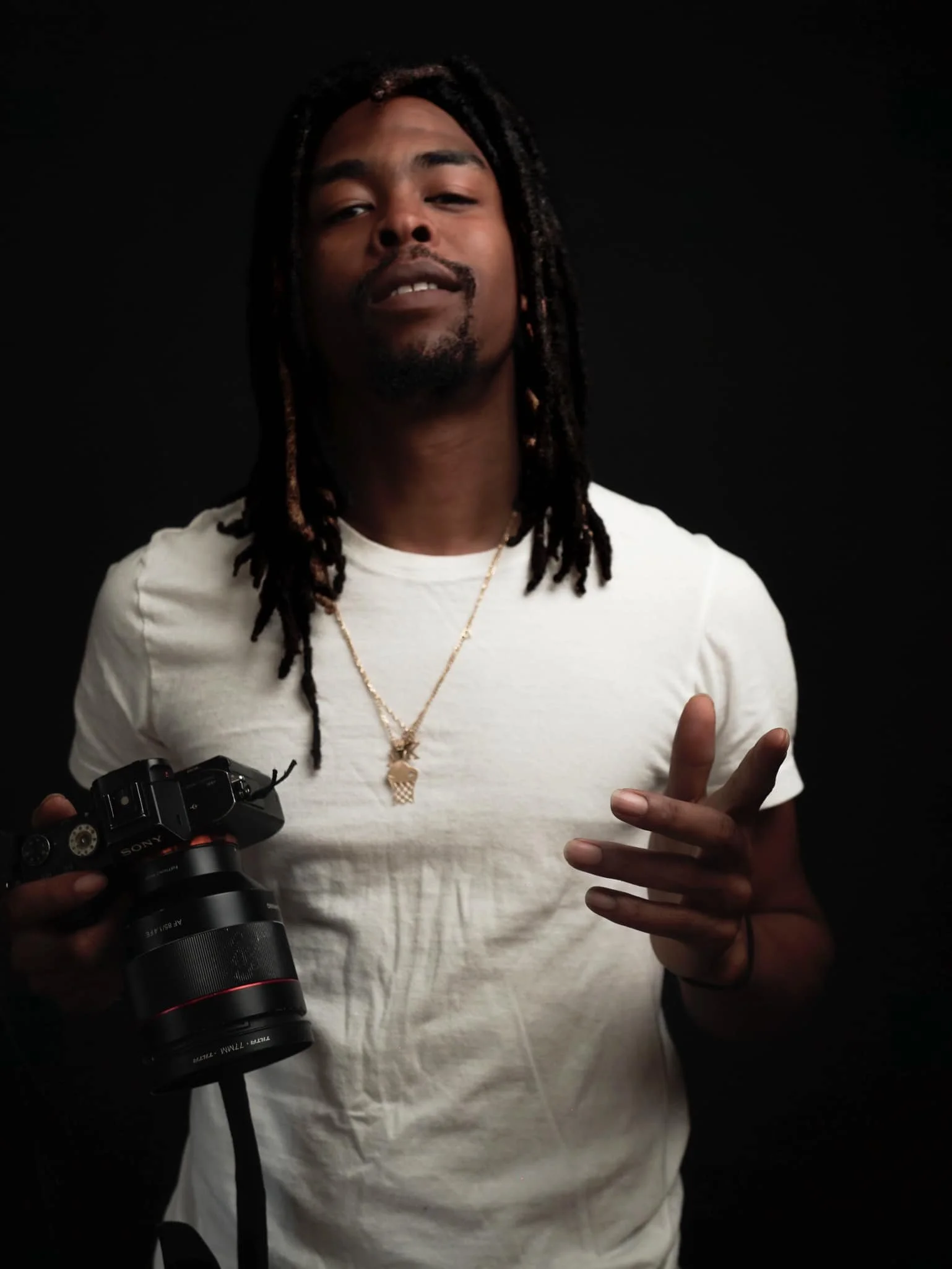 A young man with dreadlocks holding a professional camera, wearing a white t-shirt and gold jewelry, posing against a black background.