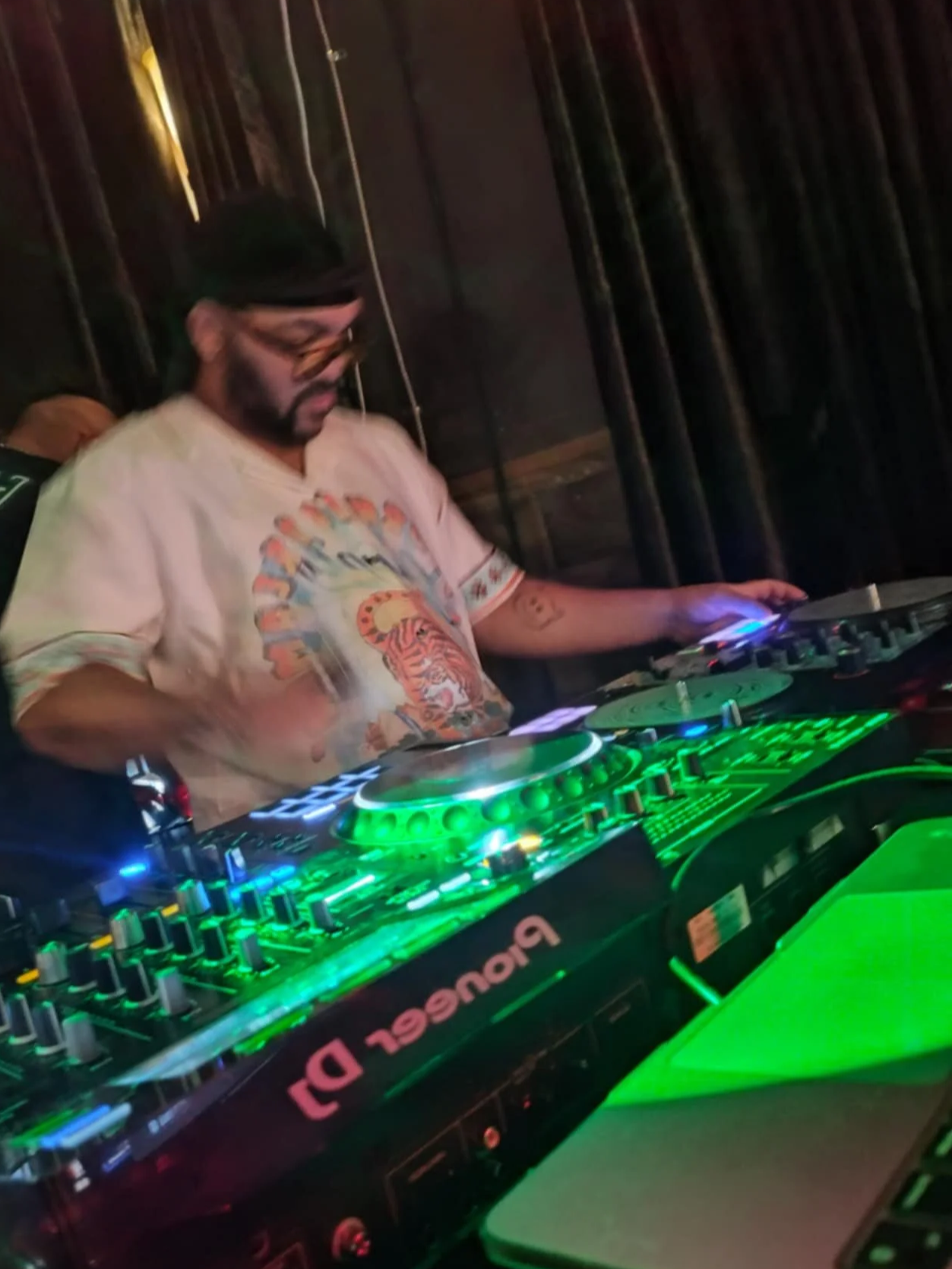 A DJ wearing glasses and a printed t-shirt, focused on his mixing console with green LED lights, performing at a dimly lit venue with dark curtains in the background.