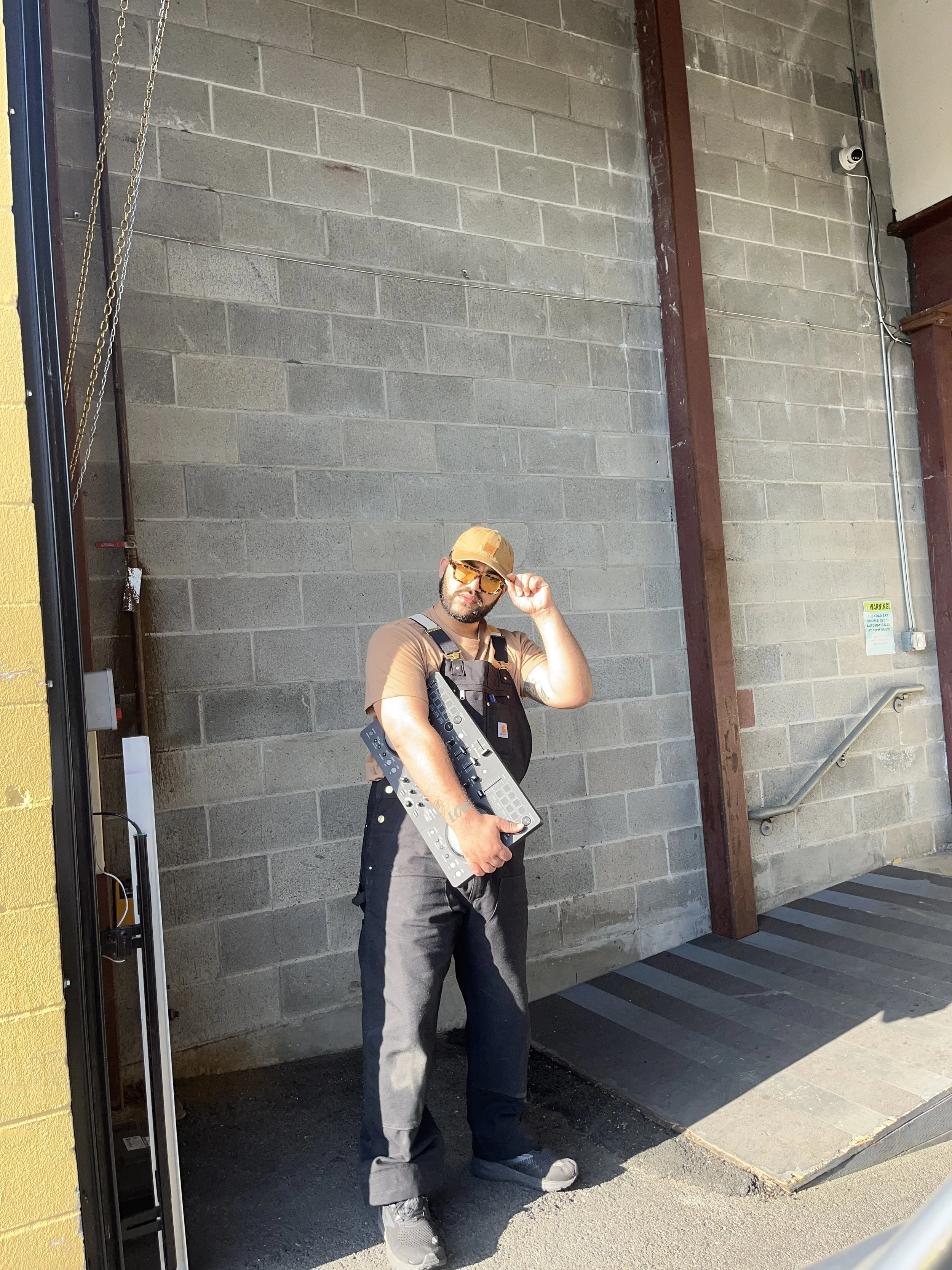A man standing outside near a brick wall, wearing sunglasses and a yellow cap, holding a MIDI keyboard controller, dressed in a brown T-shirt and black overalls.