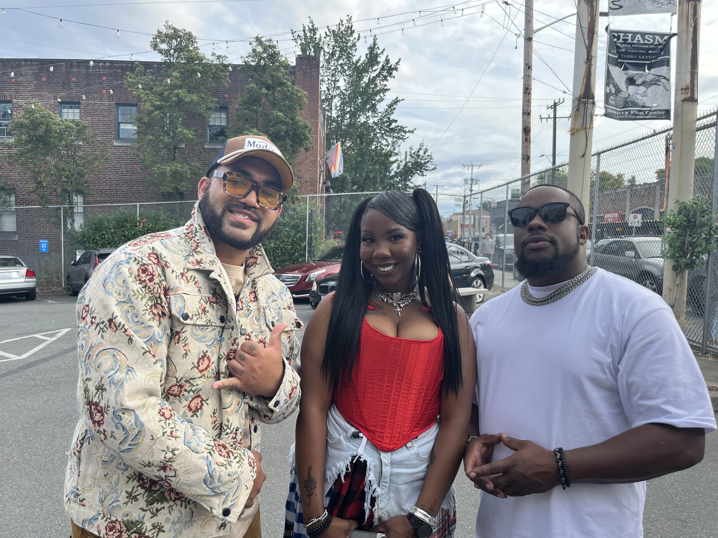 Three people standing outdoors in a parking lot. The person on the left wears a floral patterned jacket, sunglasses, and a baseball cap. The woman in the middle wears a red top, white shorts, and has long black hair. The person on the right wears a white t-shirt, sunglasses, and a chain necklace.