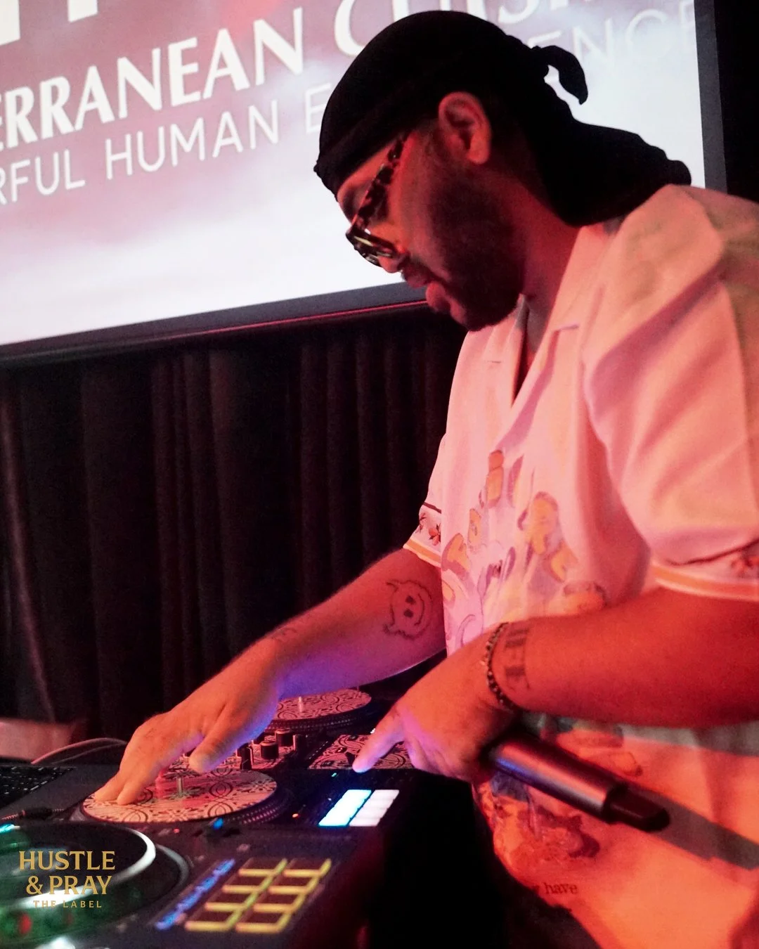 A man DJing at an event, wearing sunglasses, a black head covering, and a cream-colored shirt, with a large screen behind him displaying text.