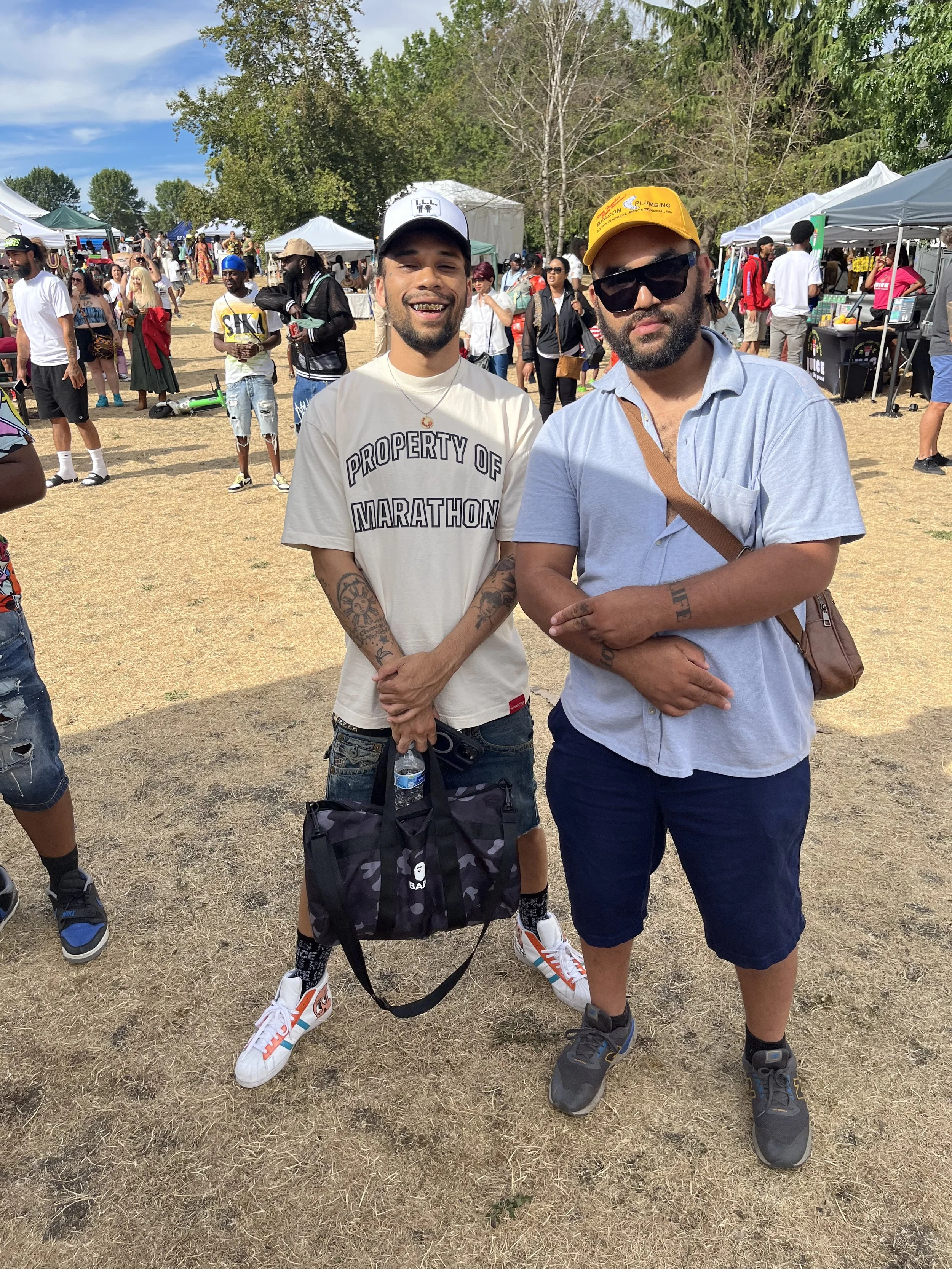 Two men standing outdoors at a festival or outdoor event, with tents and people in the background. One man is smiling and wearing a white t-shirt that says 'Property of Marathon' and holding a water bottle and a bag. The other man is wearing sunglasses, a yellow cap, and a light blue shirt.