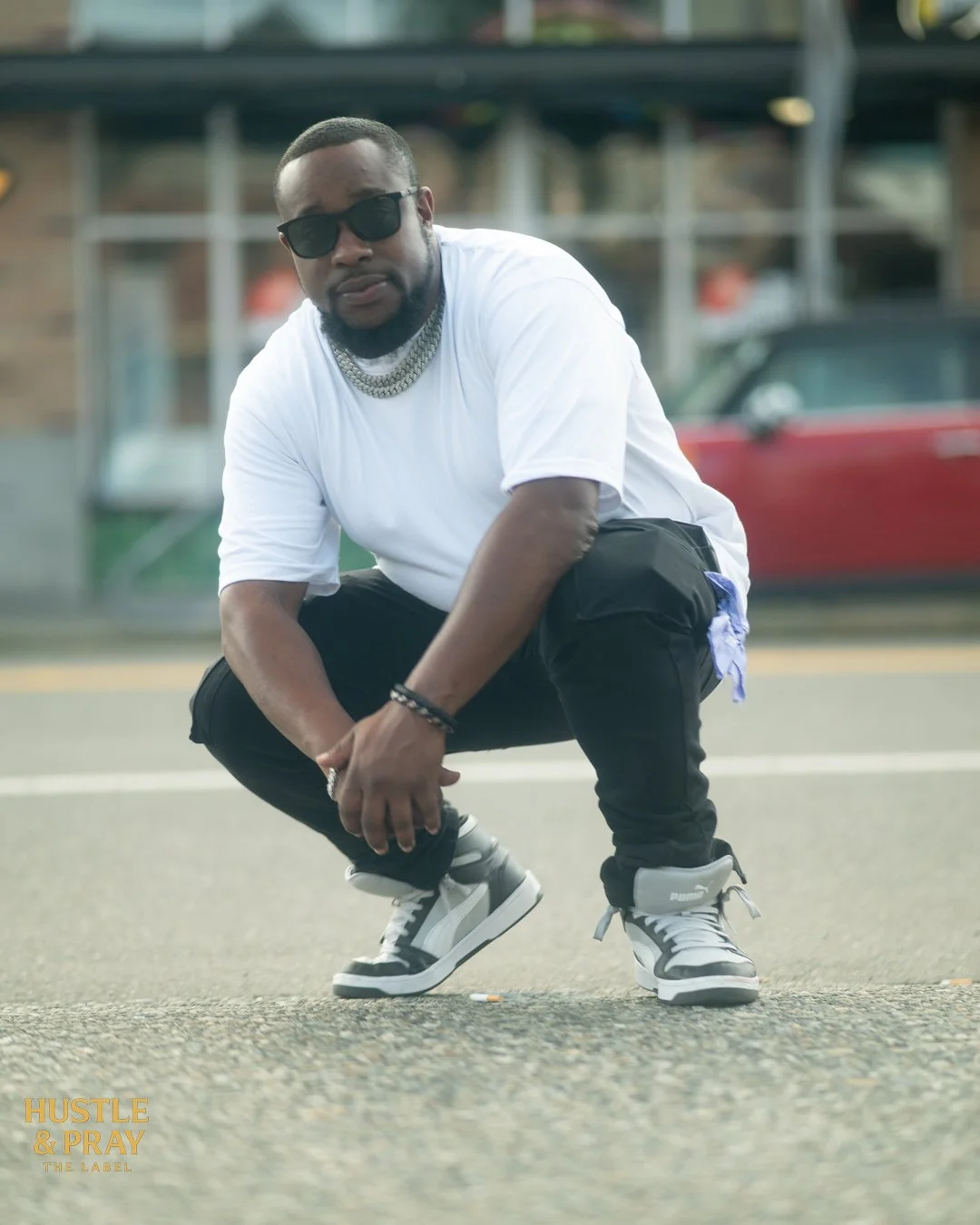 A man crouching on a street wearing sunglasses, a white t-shirt, black pants, and gray sneakers, with a gold chain and bracelets, with cars and buildings in the background.