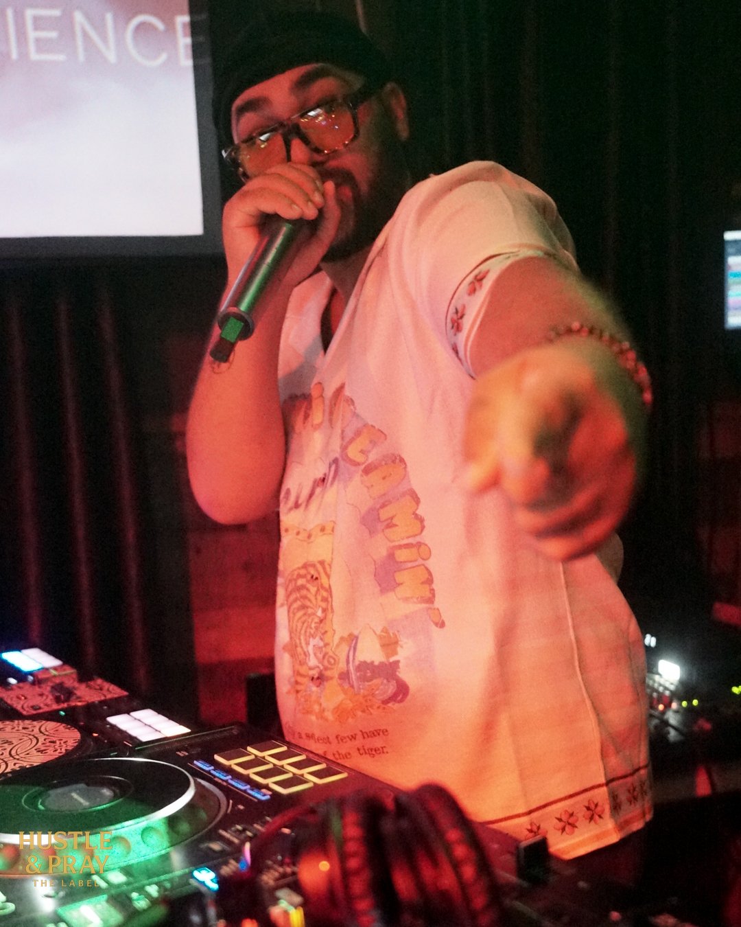 A DJ or performer with glasses, a beard, and a black cap, holding a microphone and pointing towards the camera, in a dimly lit setting with DJ equipment in front.