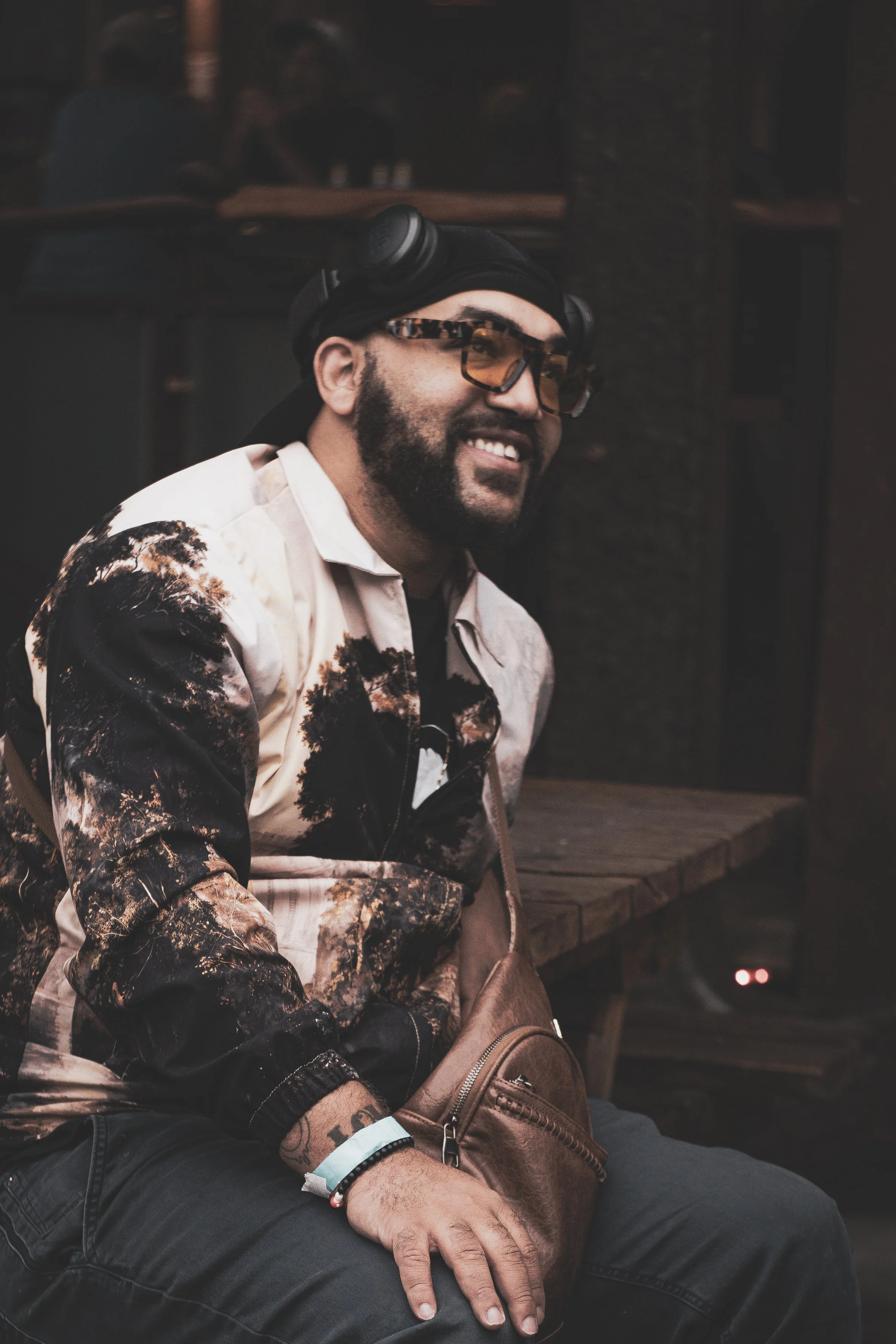 A smiling man with a beard wearing sunglasses, a black beanie, camouflage jacket, and black pants, sitting indoors with a brown crossbody bag on his lap.