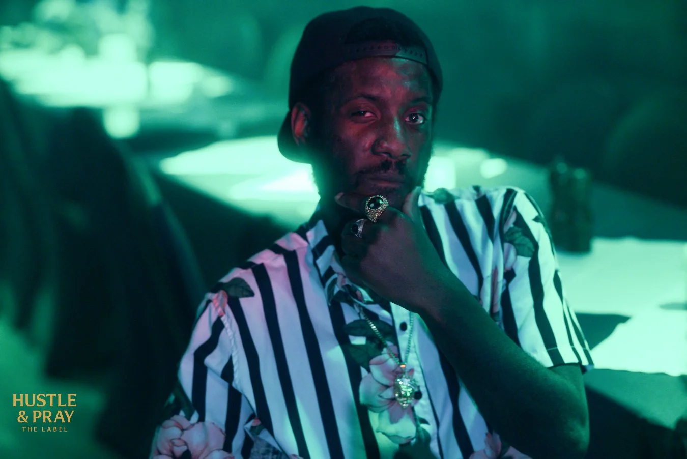 A man with dark skin wearing a black baseball cap backwards, a floral patterned shirt with black and white stripes, and jewelry including rings and a chain, is sitting in a car with neon lighting. He is posing with his hand on his chin, looking at the camera.
