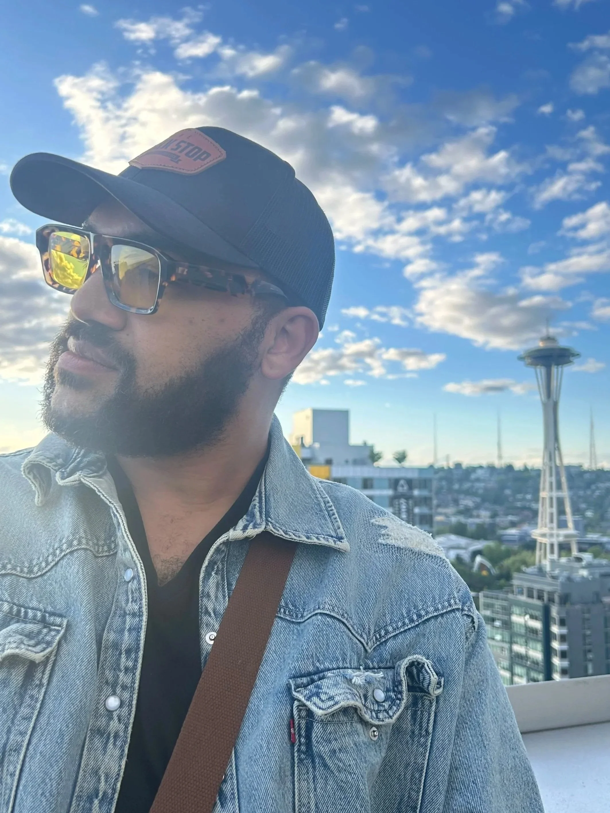 Man with a beard wearing sunglasses, a black cap, a denim jacket, and a brown strap bag, with the Space Needle and cityscape in the background under a partly cloudy sky.