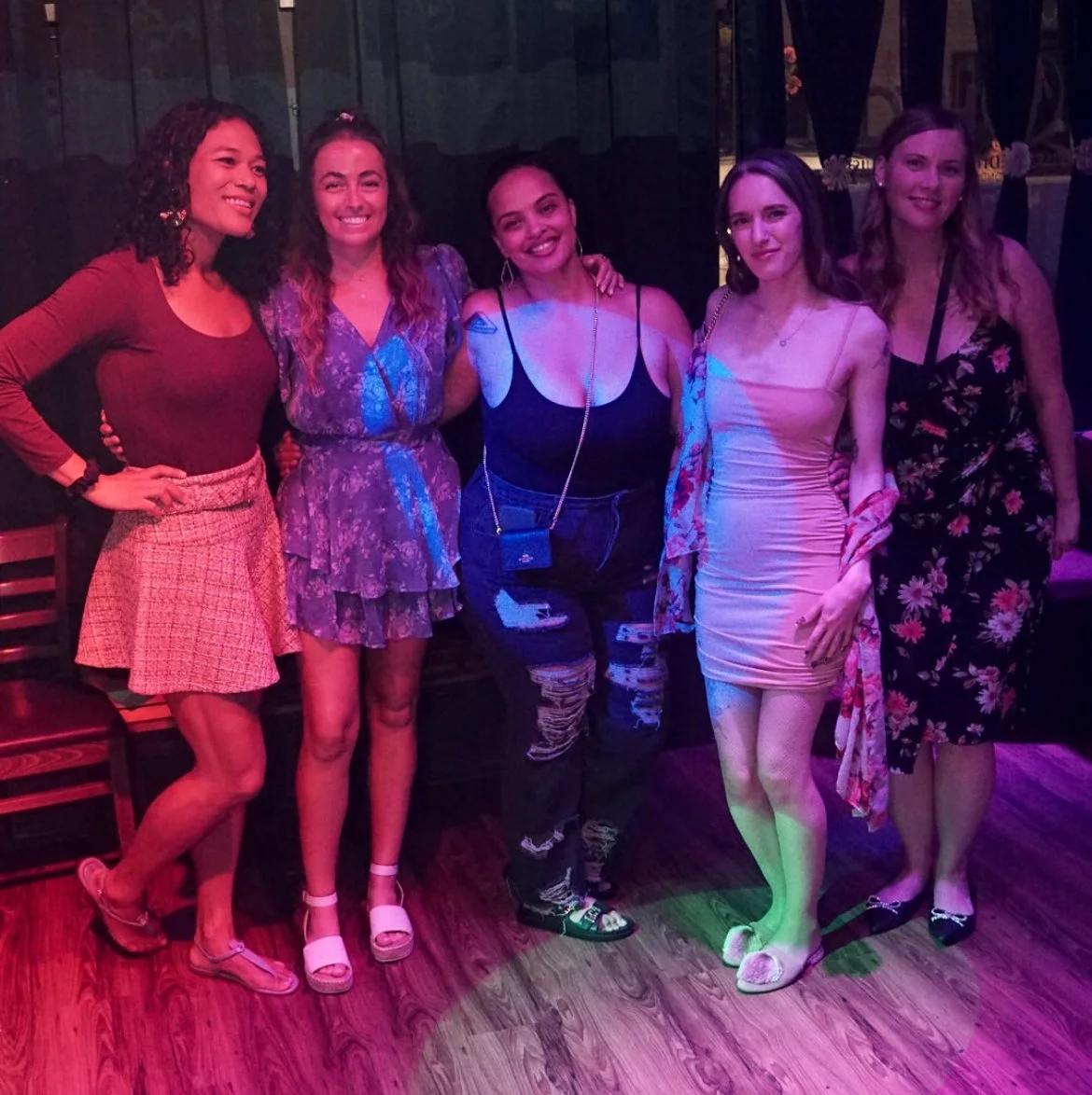 Group of five women standing together in a dimly lit venue, smiling and posing for the camera.
