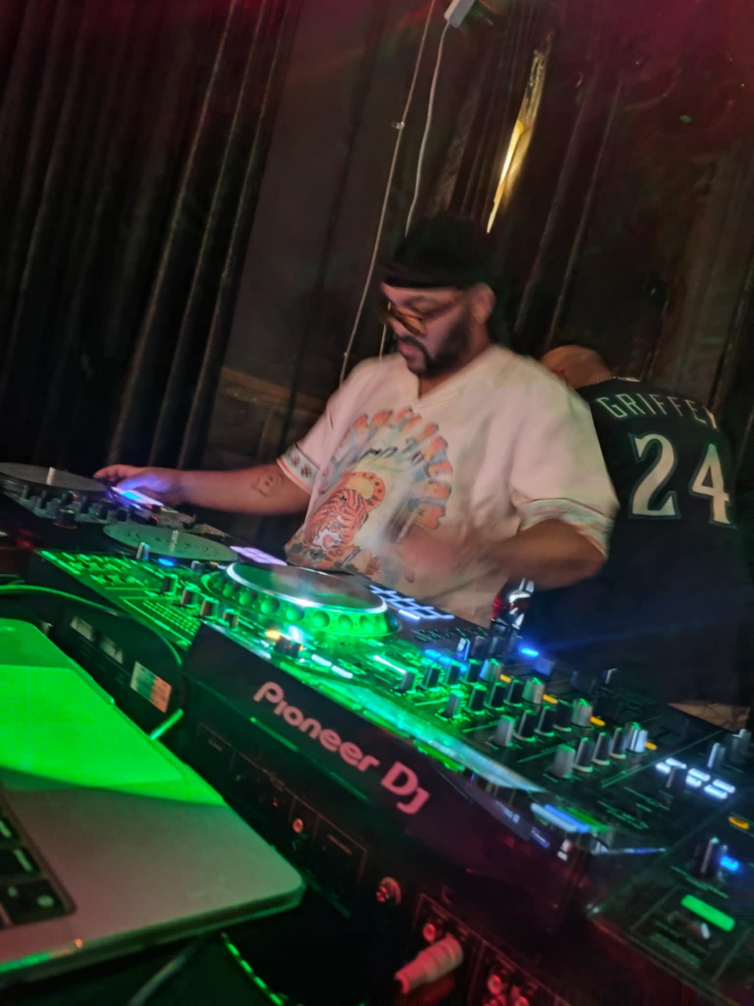 A DJ with tattoos and glasses wearing a beige shirt, standing behind a Pioneer DJ controller with colorful lighting, in a dark environment, possibly a nightclub or music venue.