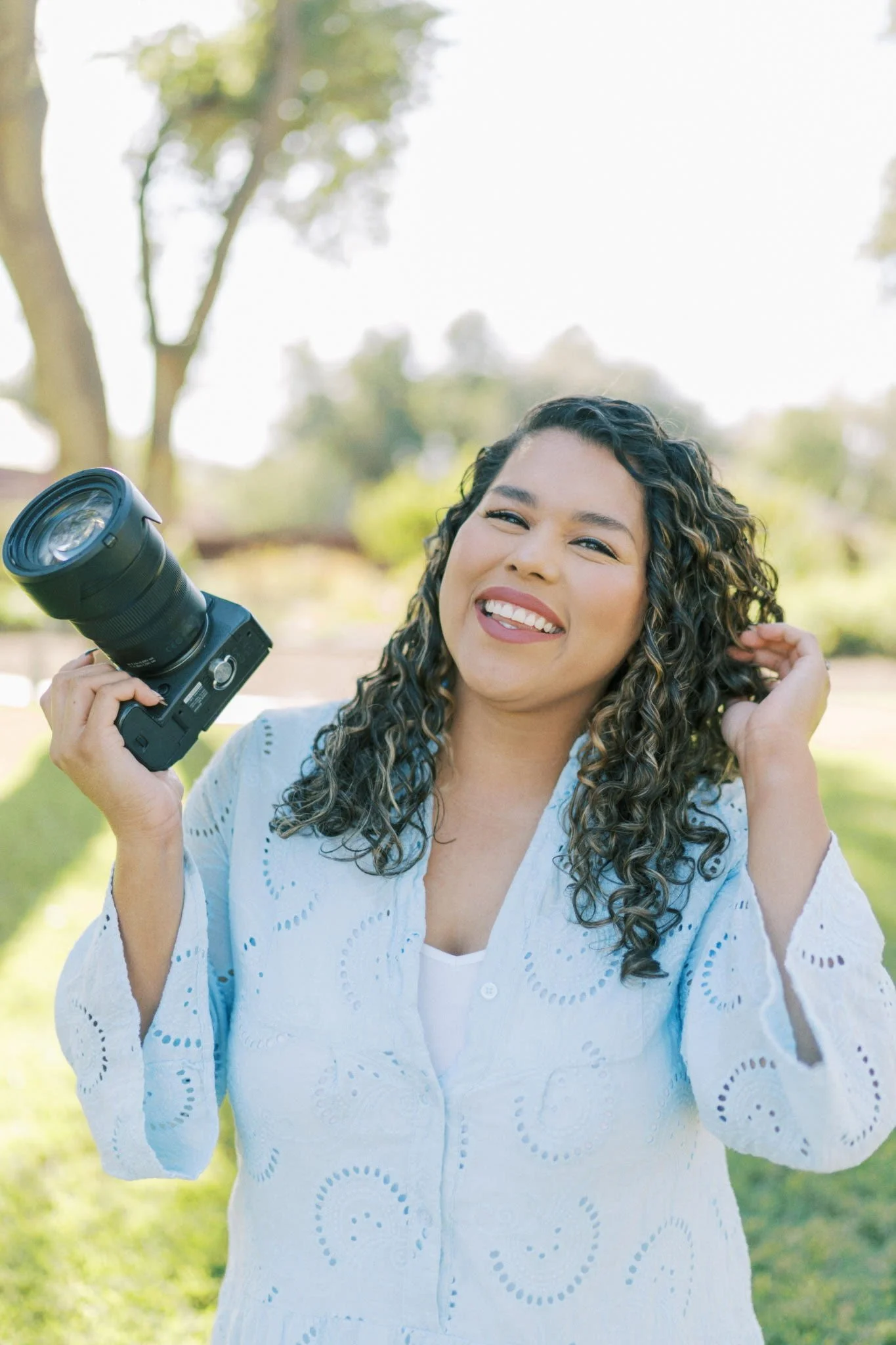 Smiling woman with curly hair outdoors, holding a camera in her right hand, wearing a white blouse with eyelet details, trees and bright sky in the background.