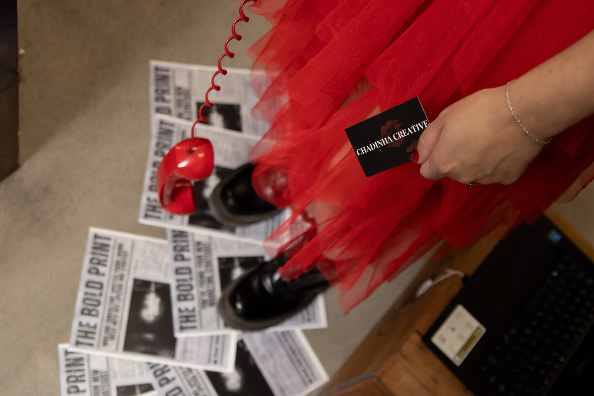 Owner's hand holding a business card that says 'Chadinha Creative' next to a vintage red toy phone with a curly cord. In the background, there are newspapers on the floor.