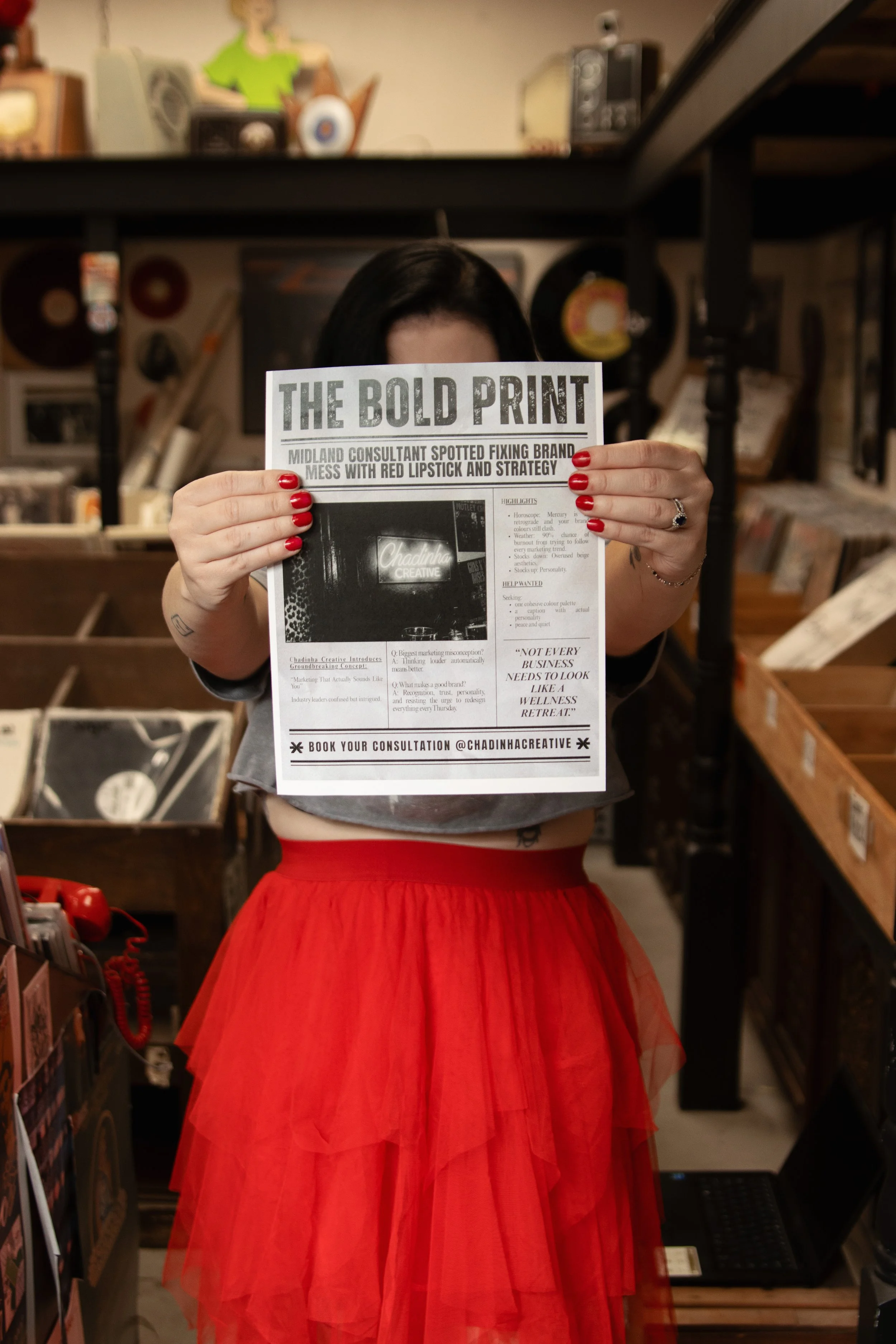 A woman wearing a red tulle skirt holding a newspaper in front of her face, with a background of shelves filled with vinyl records and old-style radios.