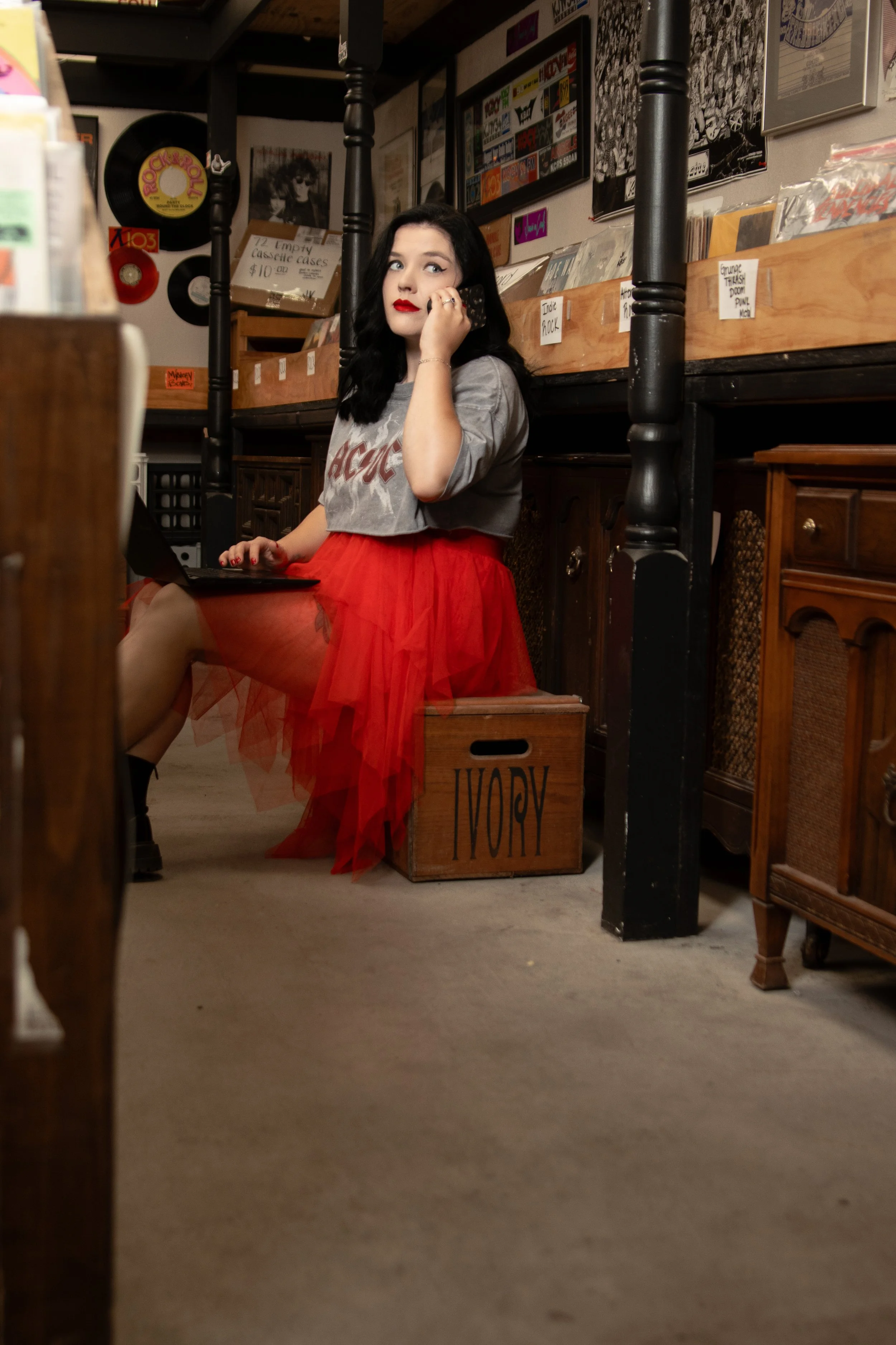 Owner with black hair, red lipstick, and vintage-style makeup, sitting on a wooden crate, wearing a gray AC/DC t-shirt and a red tulle skirt, in a room decorated with vintage music albums and posters.