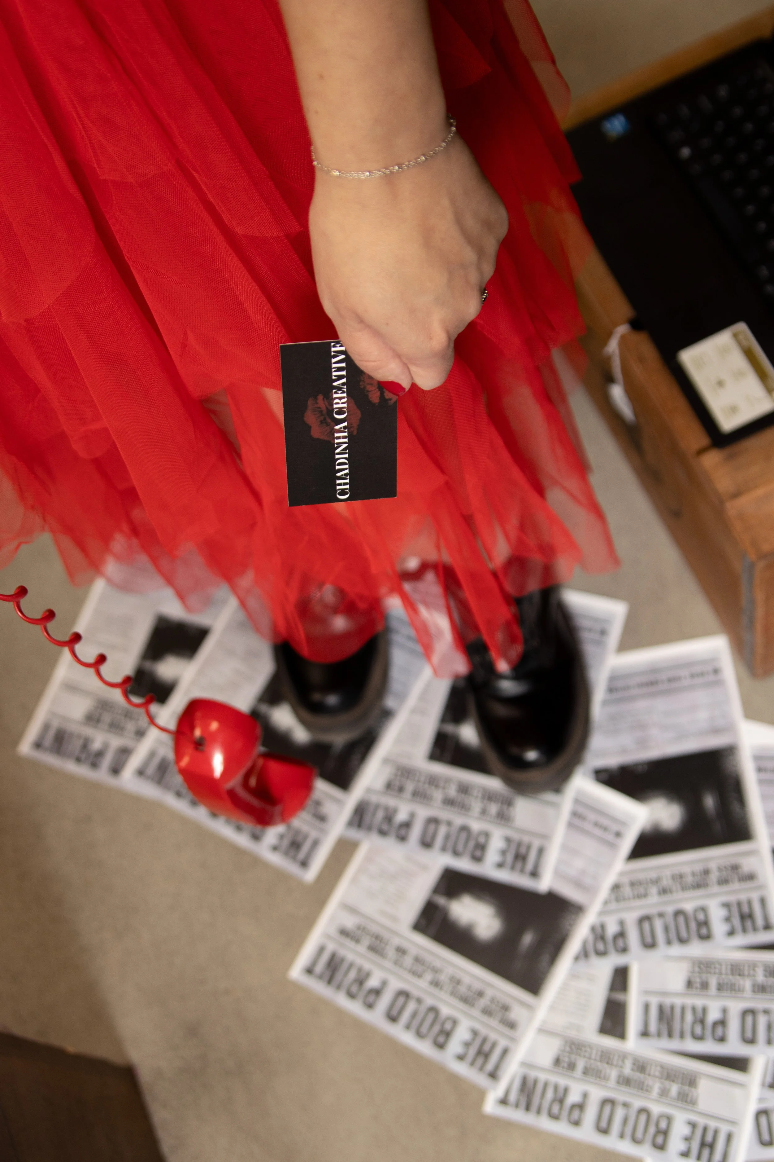 A person's hand wearing a silver bracelet and a ring, holding a black card labeled 'CHADINIA CREATIVE.' The person is dressed in a red, tulle or chiffon garment. In the background, there's a red corded telephone, some papers, and a wooden item, potentially a shelf or desk, with a black keyboard and what appears to be a box or book.