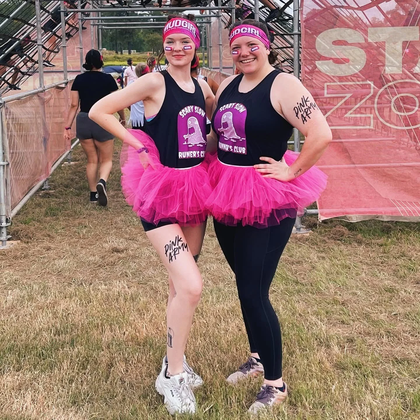 Two women at an outdoor event dressed in athletic wear and pink tutus, wearing matching headbands and face paint supporting the Pink Army, with a grassy field and event structure in the background.