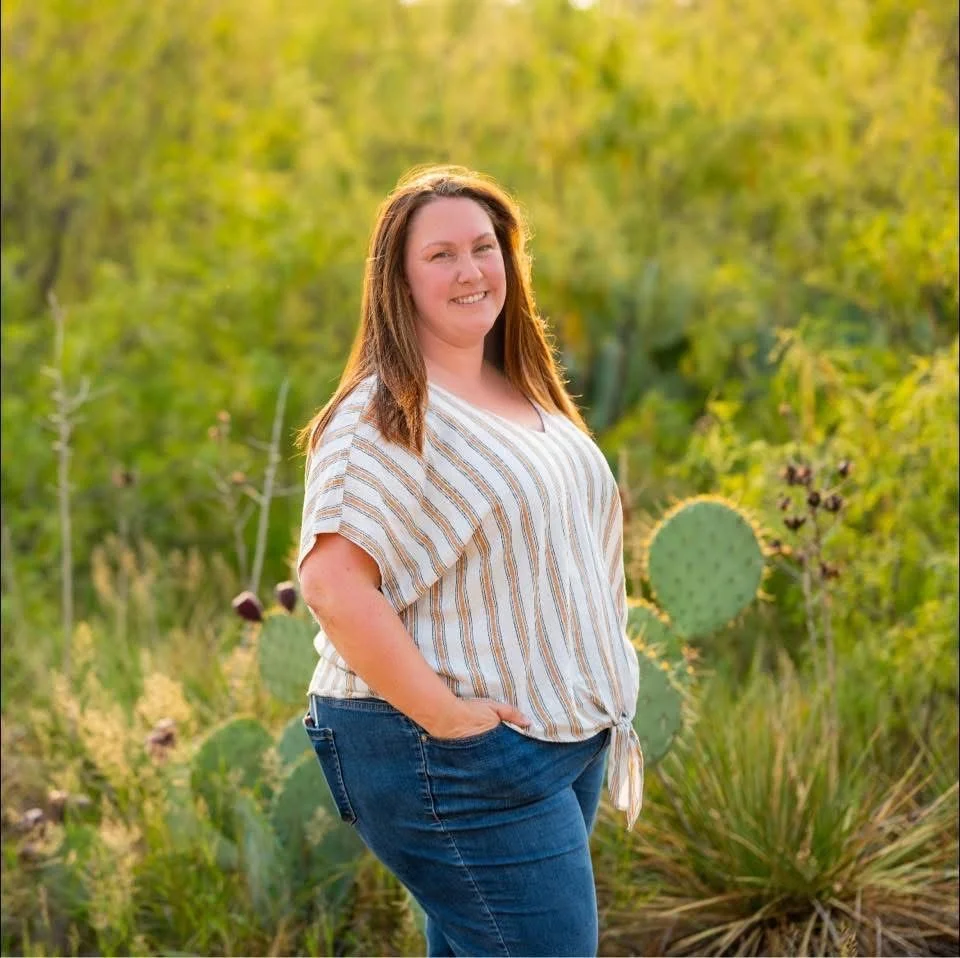 A smiling woman standing outdoors among green desert plants, including prickly pear cacti, during daytime.