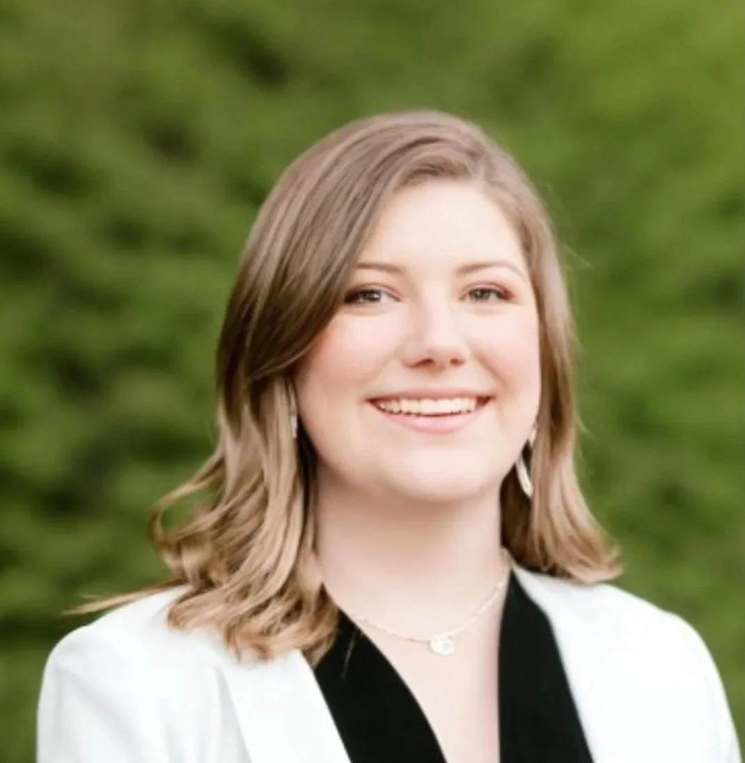 A young woman with shoulder-length light brown hair, smiling, wearing a white blazer with black lapels and a gold necklace, outdoors with a green leafy background.