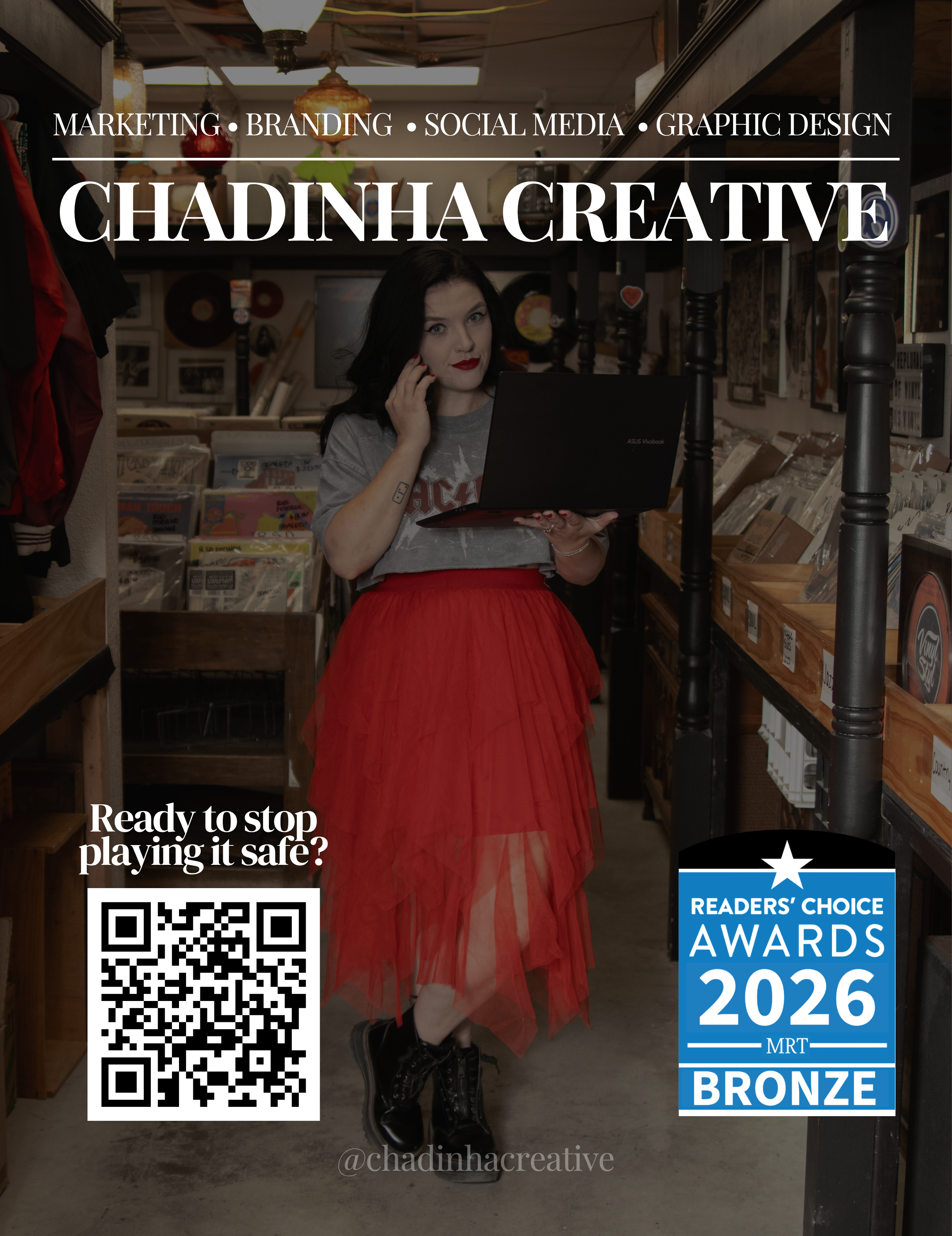A woman with black hair, red lipstick, wearing a gray T-shirt, a red tulle skirt, and black lace-up boots standing in a store aisle. She is holding a black laptop and talking on her phone, with store shelves filled with books and records behind her.