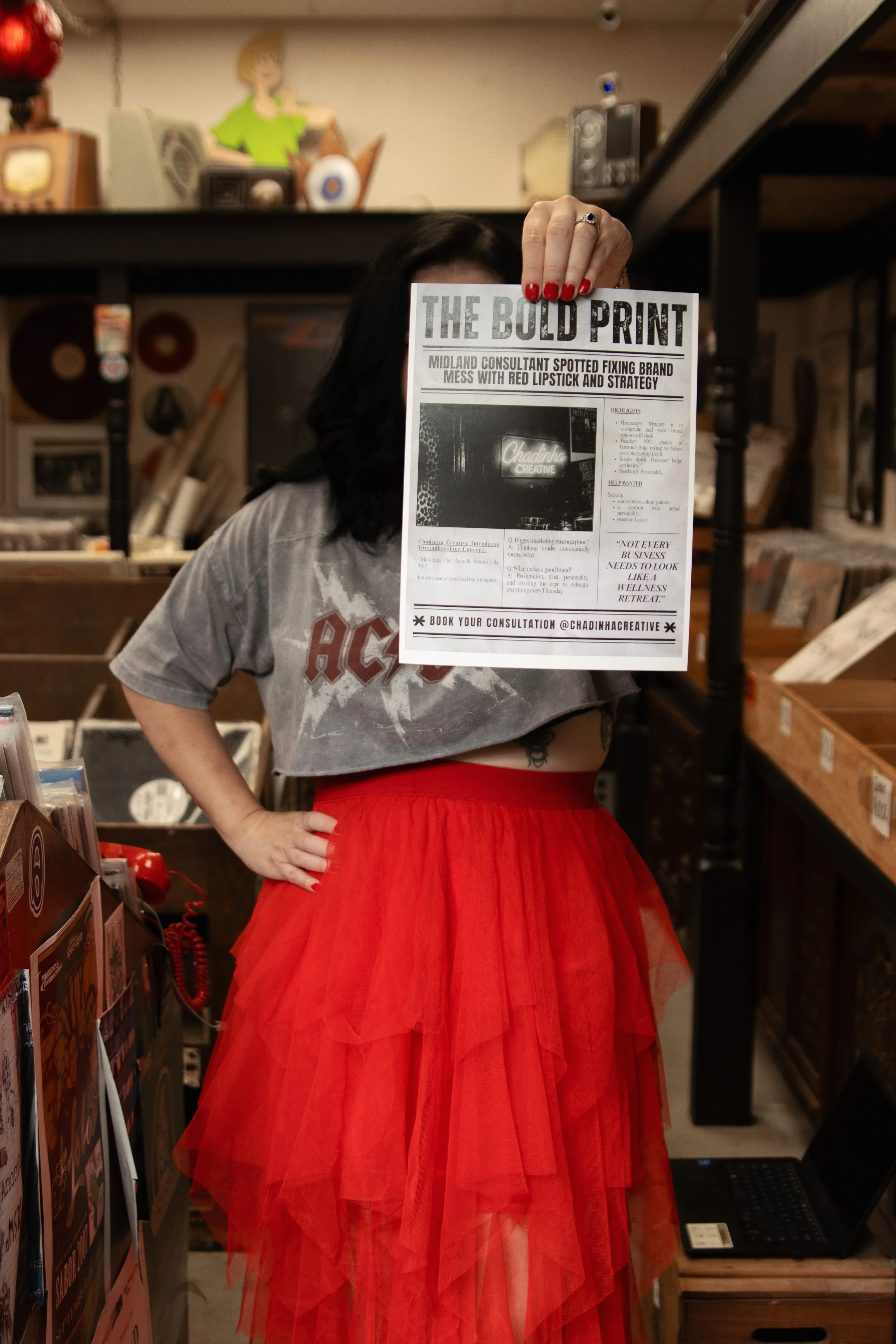 Owner holding a black-and-white newspaper in front of their face, wearing a gray AC/DC t-shirt and a red tulle skirt, in a store with vinyl records and vintage decor in the background.