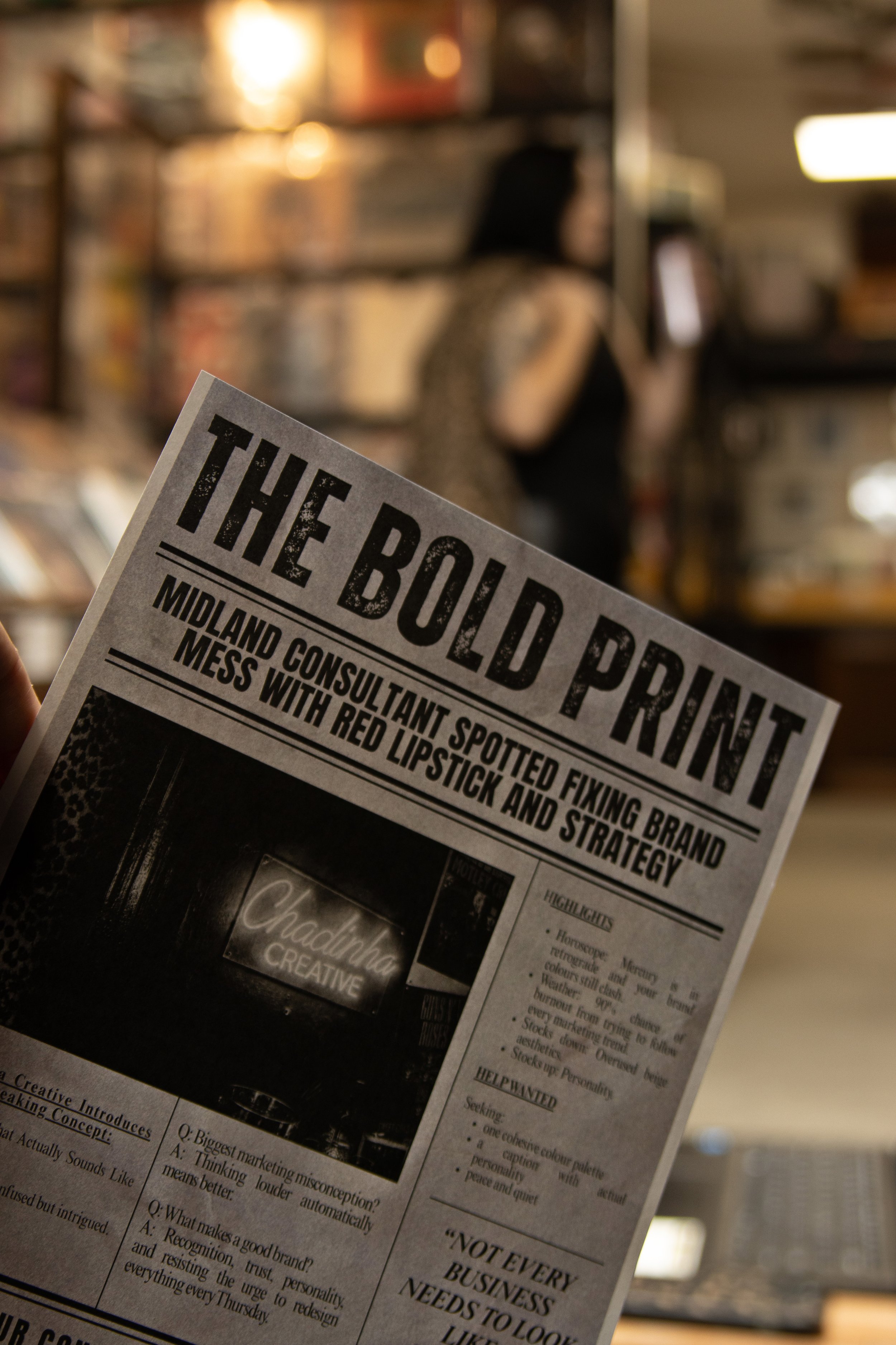 A person holding a newspaper titled 'The Builder' in a bookstore or library. In the background, a woman is seen browsing shelves of books.