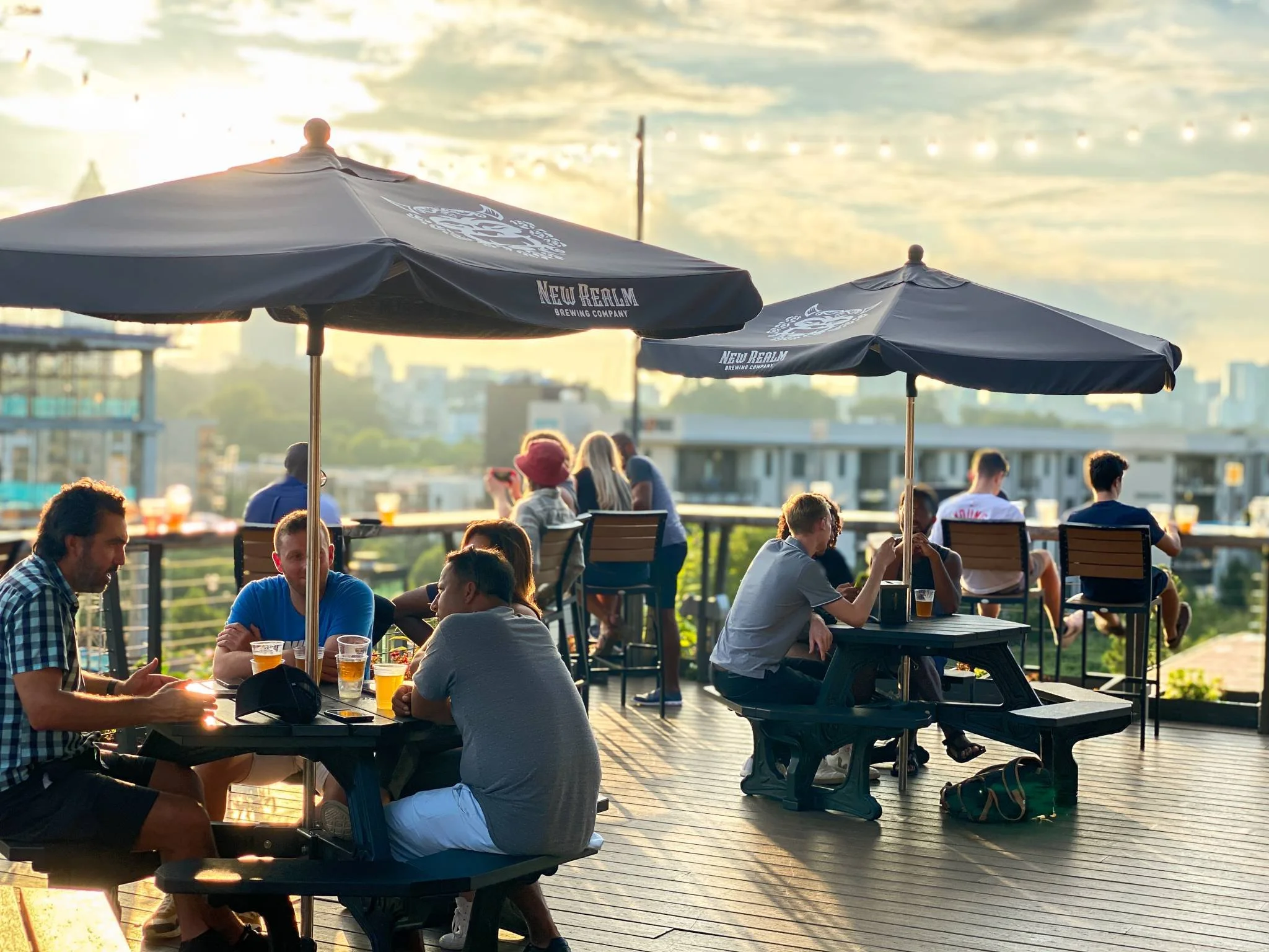 People sitting at outdoor tables with umbrellas on a rooftop, enjoying drinks during sunset with city buildings in the background.