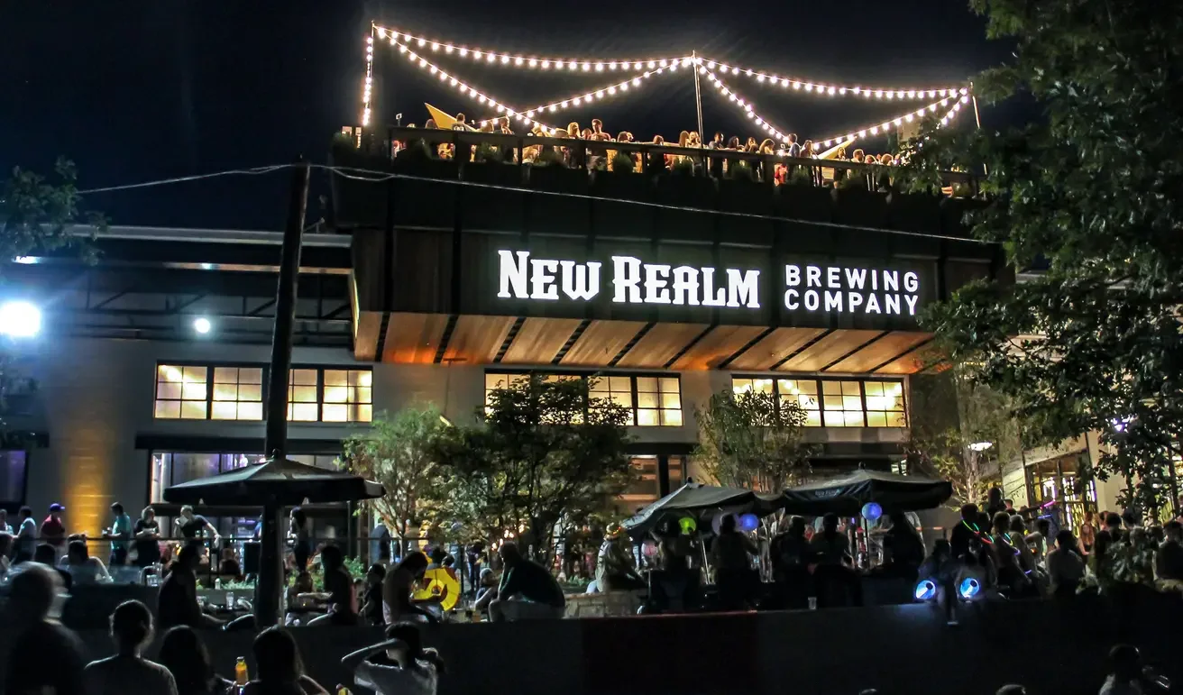 Night view of New Realm Brewing Company with a crowd outside and rooftop patio illuminated with string lights.