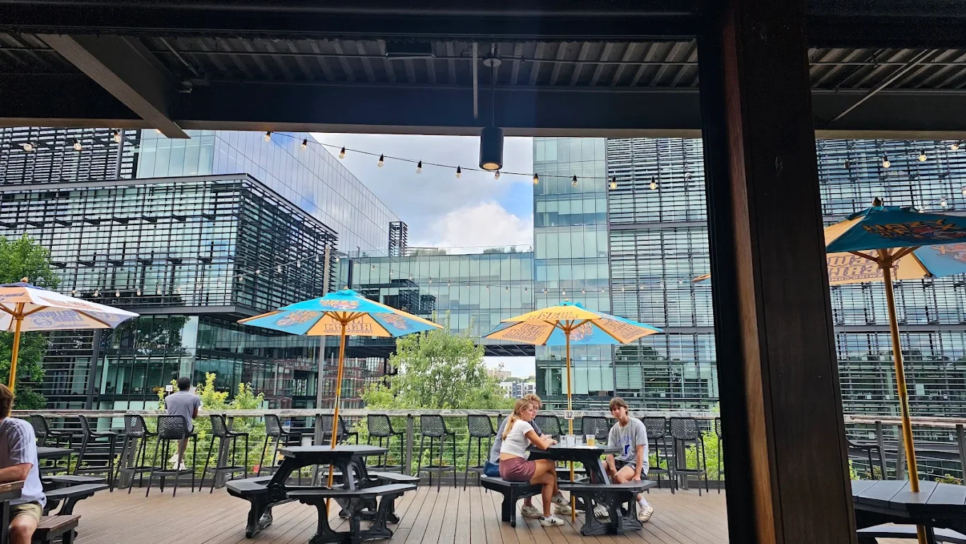 Outdoor patio with several people sitting at tables under umbrellas, modern glass buildings in the background.