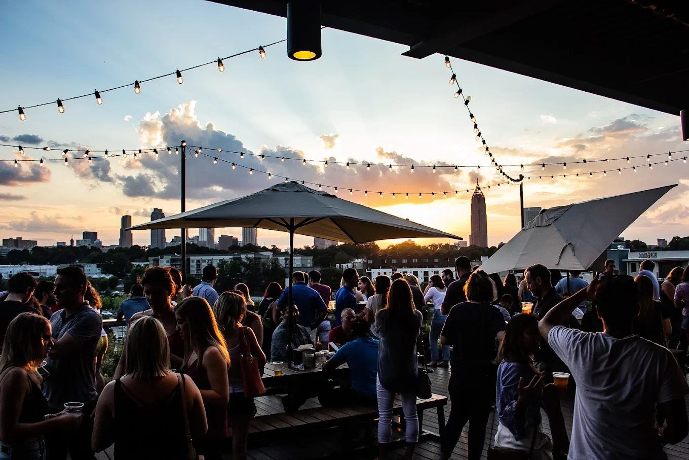 People gathered at a rooftop party during sunset, with string lights and umbrellas, overlooking a city skyline.