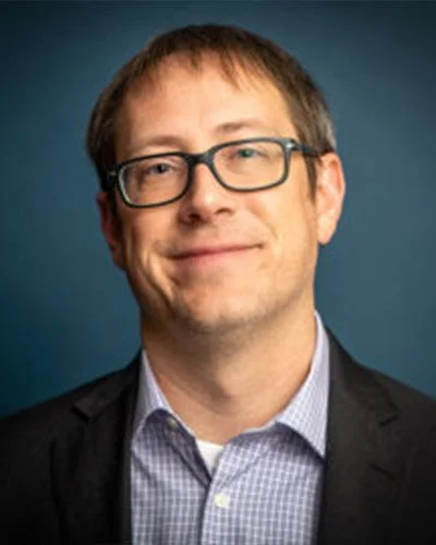 Portrait of a man with glasses, short brown hair, wearing a suit jacket and collared shirt, smiling against a blue background.