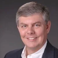 Headshot of a man with gray hair dressed in a dark suit and white shirt, smiling against a gray background.