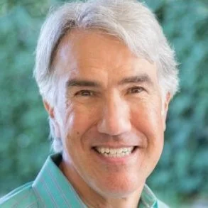 A smiling middle-aged man with gray hair outdoors with green foliage in the background.