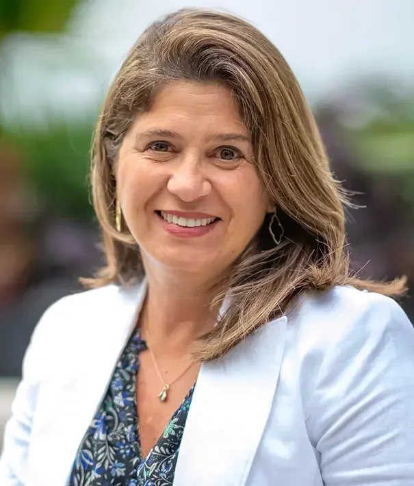 A smiling woman with shoulder-length light brown hair, wearing a white blazer, a blue patterned blouse, earrings, and a necklace, outdoors with a blurred background.