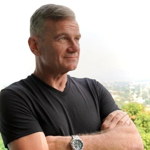 A middle-aged man with short gray hair wearing a black t-shirt and a silver wristwatch over his crossed arms, standing outdoors with a blurred city skyline and greenery in the background.
