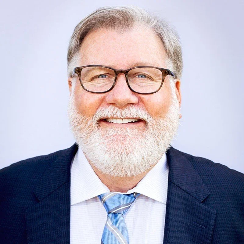 A smiling older man with gray hair and a beard, wearing glasses, a dark suit, white shirt, and a light blue striped tie.