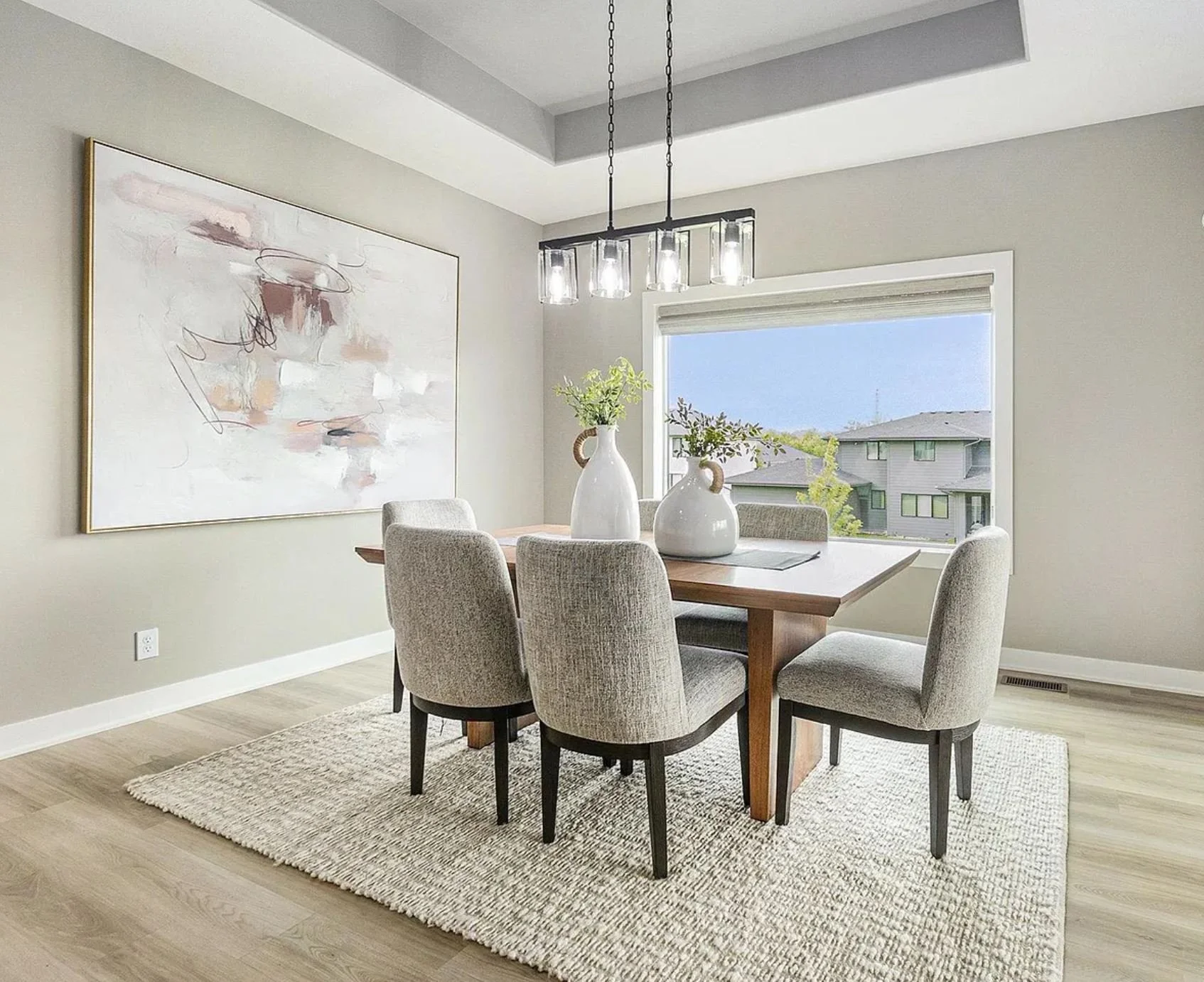 A modern dining room with six beige upholstered chairs surrounding a wooden rectangular table, two white vases with green foliage on top, a large abstract artwork on the wall, and a window with a view of neighboring houses and trees.