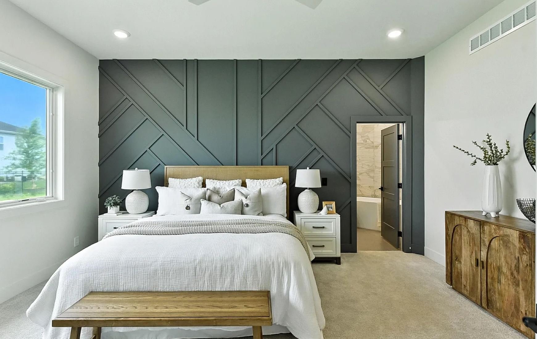 Bedroom with a gray accent wall, white bedding, and matching white nightstands with lamps. Large window on the left, wooden dresser on the right, and a doorway leading to a bathroom.