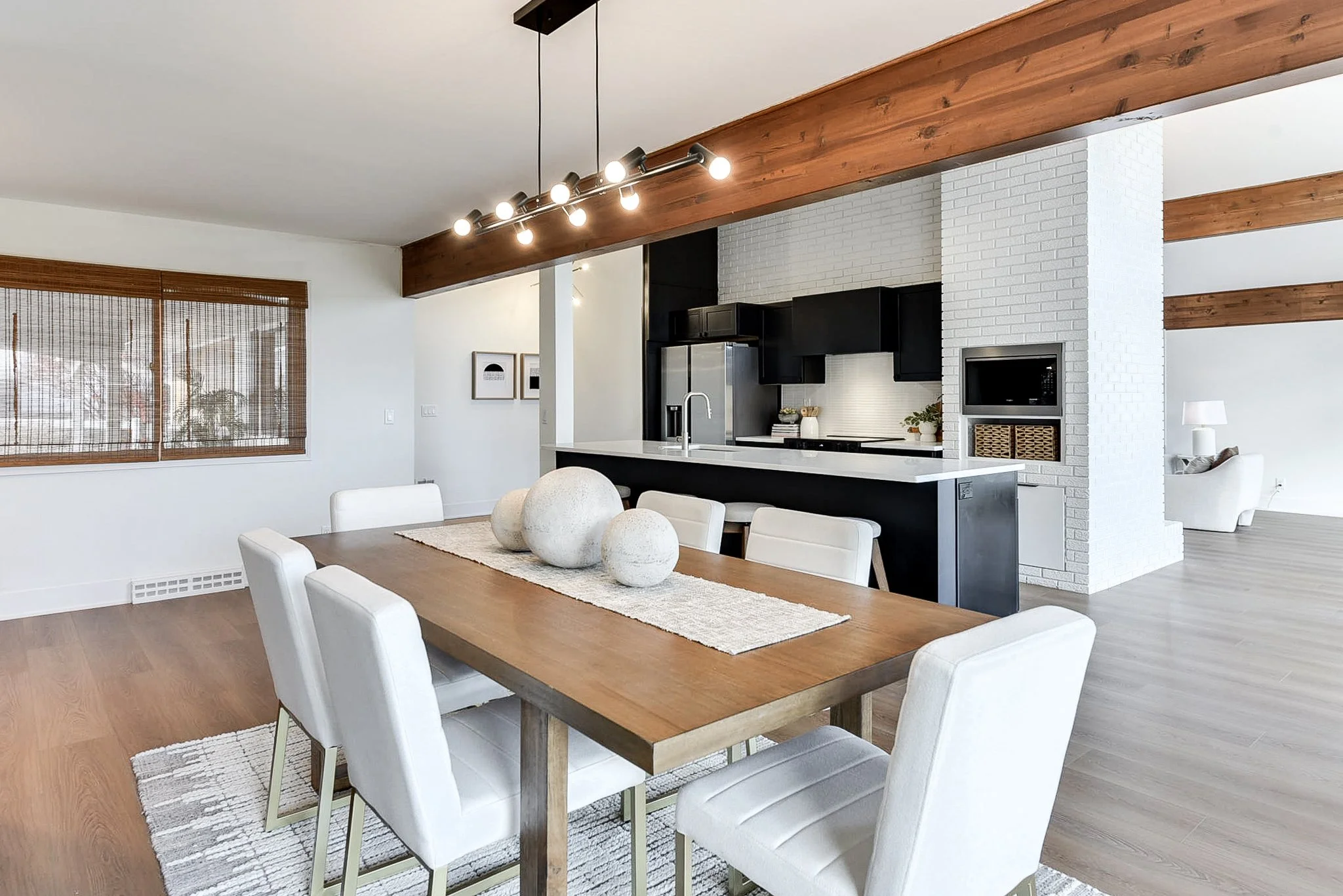 Modern open-concept kitchen and dining area with a wooden table, white chairs, black cabinets, white brick wall, and ceiling beams.