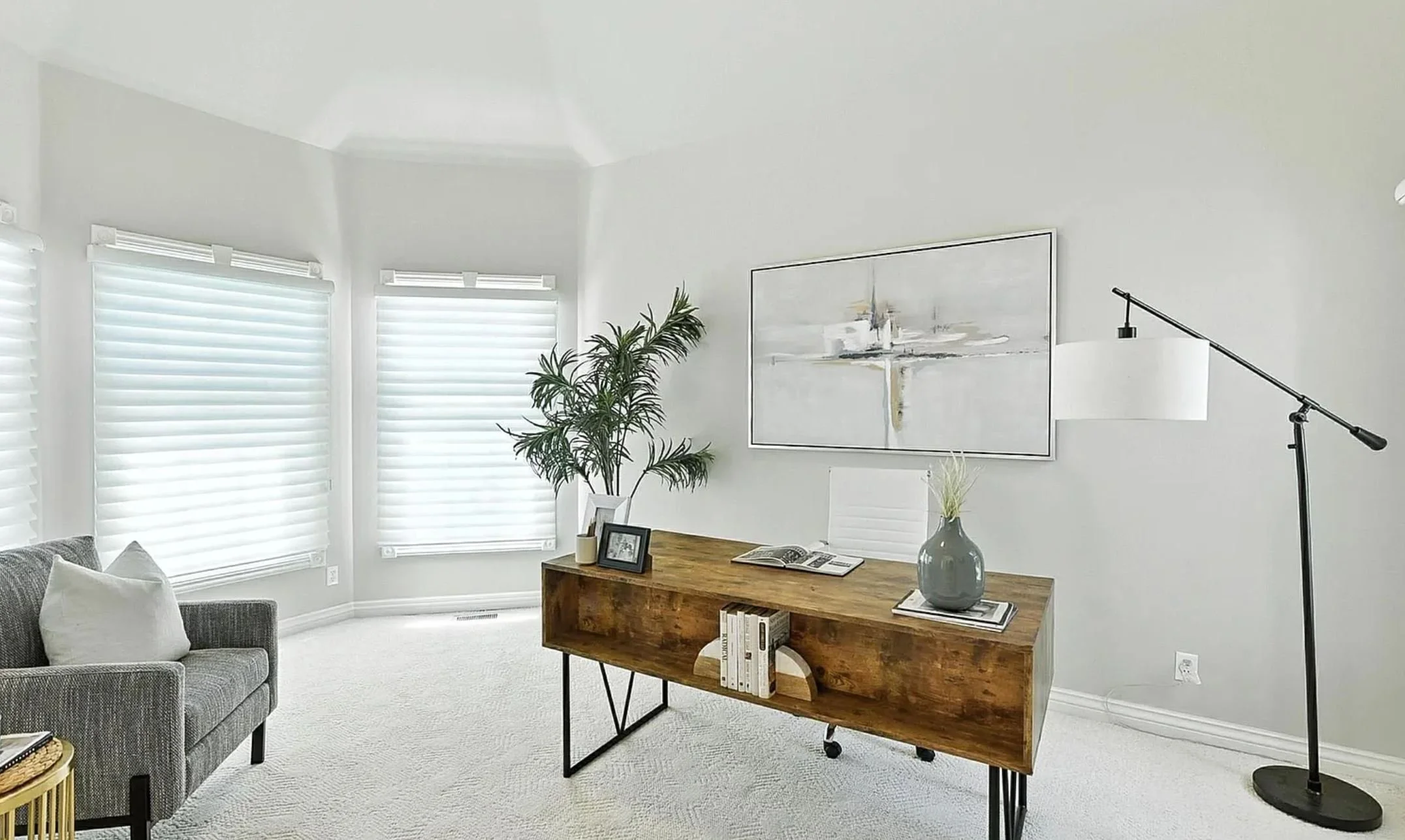 Modern office with large windows, a wooden desk, a white chair, abstract wall art, a tall green plant, a gray vase with decorative grass, and a black floor lamp.
