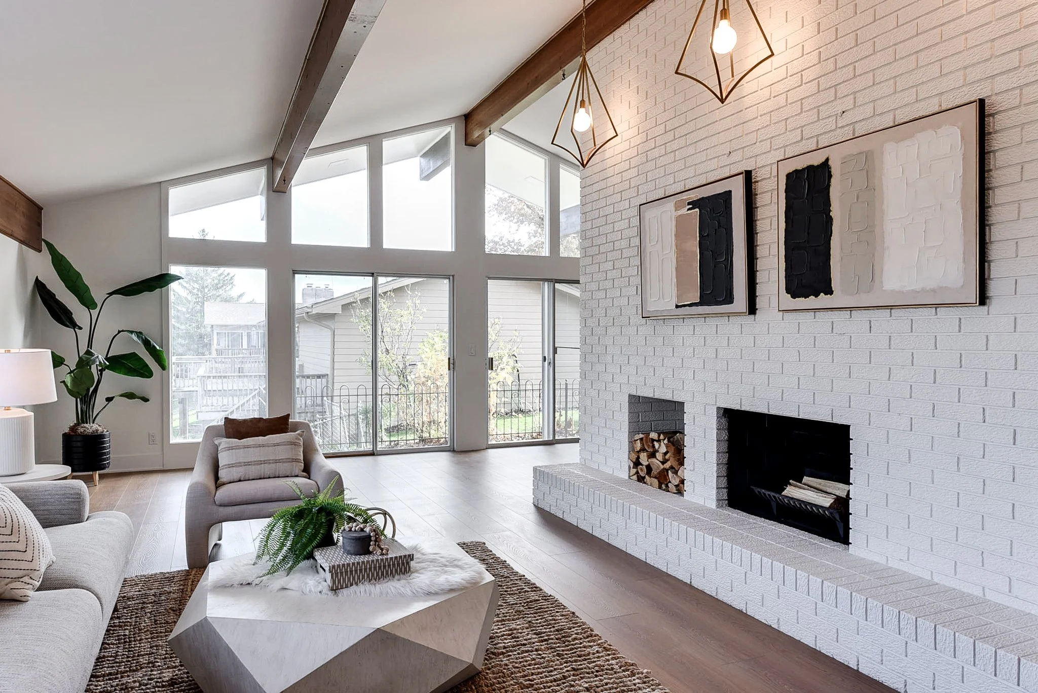 Living room with white brick fireplace, abstract wall art, large windows, and modern lighting fixtures, with wooden beams on the ceiling and a plant near the window.