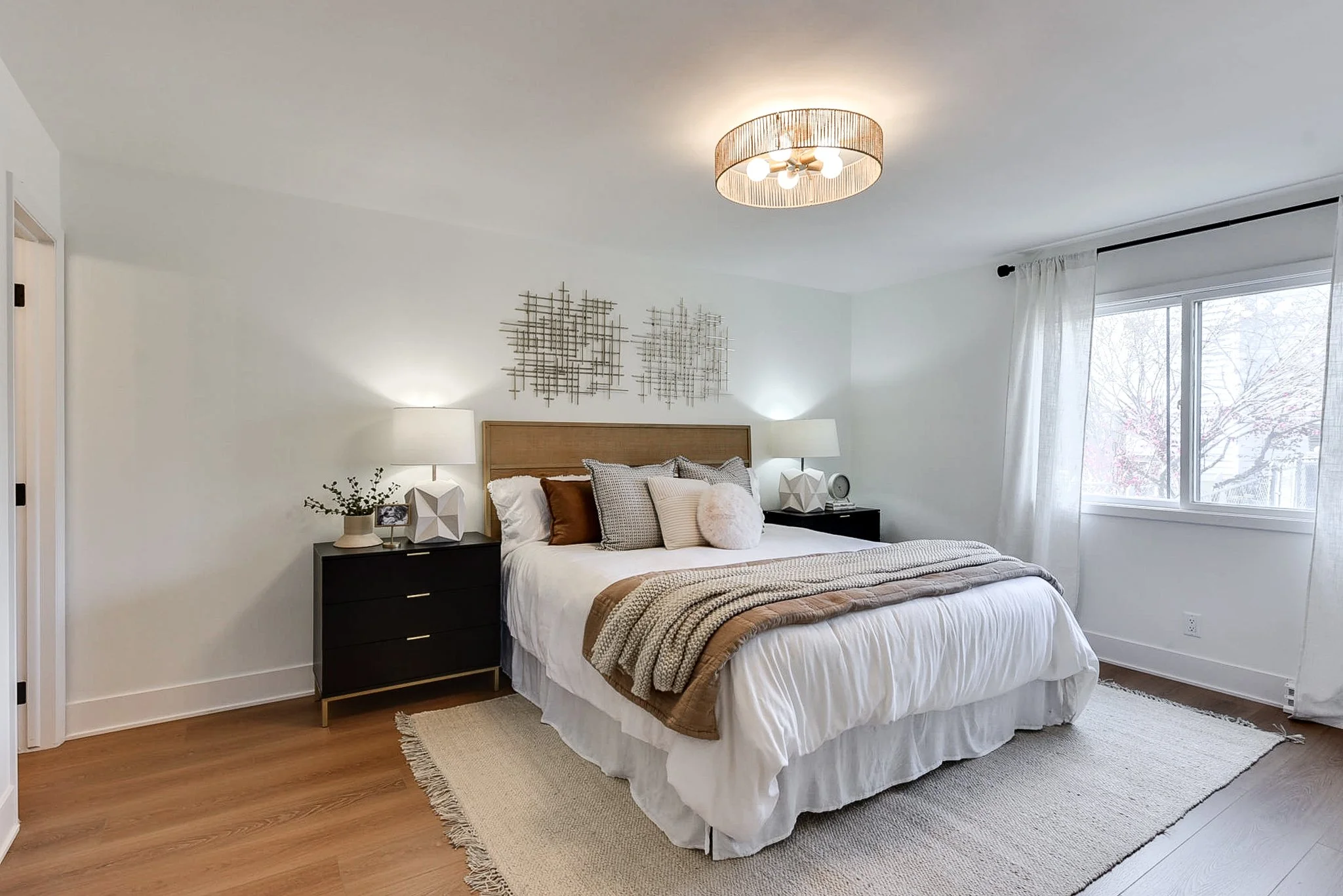 A bedroom with a neatly made bed, black nightstands with lamps, modern wall art, and a window with white curtains, hardwood floors, and a light-colored area rug.