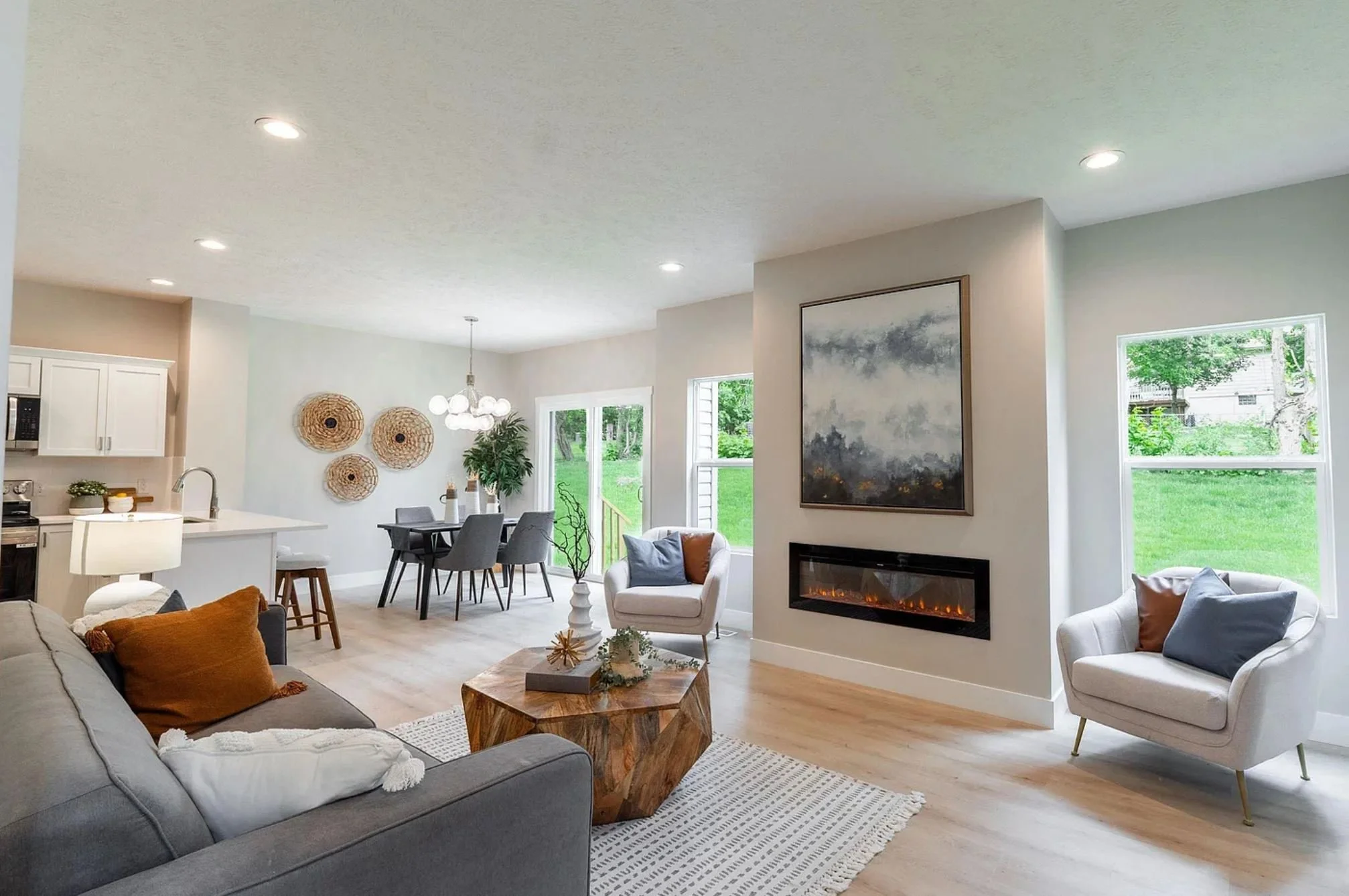 Spacious living room with a gray sofa, white armchair, wooden coffee table, and a built-in fireplace on a light-colored wall. Large windows reveal a green backyard, and the room features modern decor and recessed ceiling lights.