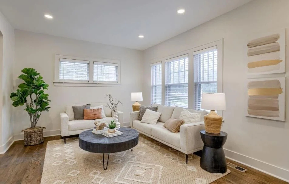 Bright living room with white sofas, neutral throw pillows, a round black coffee table, a tall green plant in a wicker basket, and two table lamps on black and beige side tables. Large windows with white blinds and abstract wall art decorate the spac