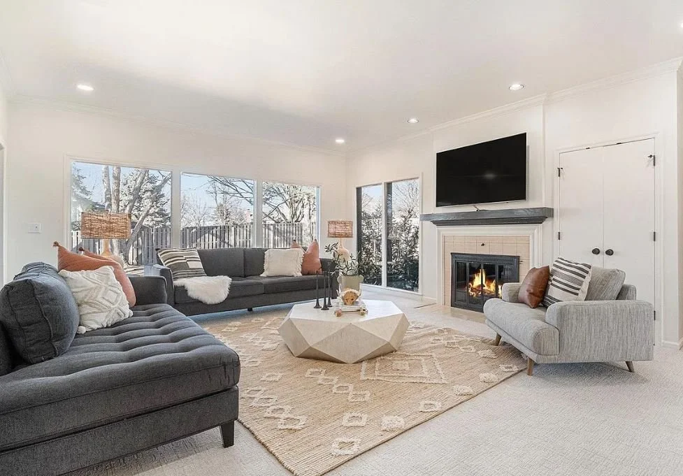 Living room with large windows, gray sofas, a white armchair, a geometric coffee table, a rug, a fireplace with a TV above, and decorative pillows.