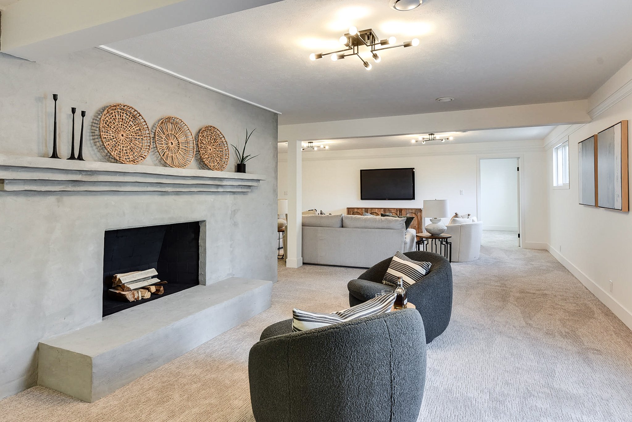 Living room with a concrete fireplace, modern chandelier, beige and gray furniture, wall-mounted TV, and minimalistic decor.