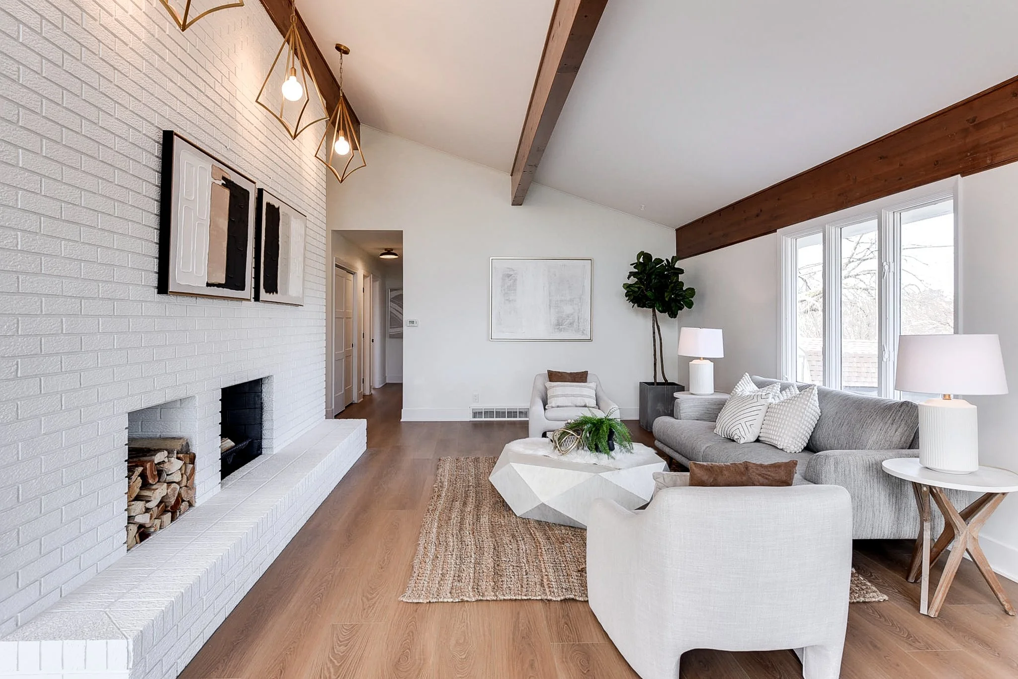 Bright living room with white brick fireplace, modern art above, gray and white sofas, wooden side tables with lamps, large windows, and a tree visible outside. There is a textured area rug and a geometric coffee table with plants.