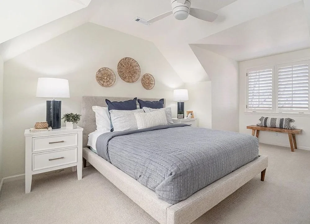 A bedroom with a neutral color palette, including a bed with gray and navy bedding, white nightstands with lamps, a woven wall art above the headboard, a window with white shutters, and a small wooden bench with a rolled pillow.