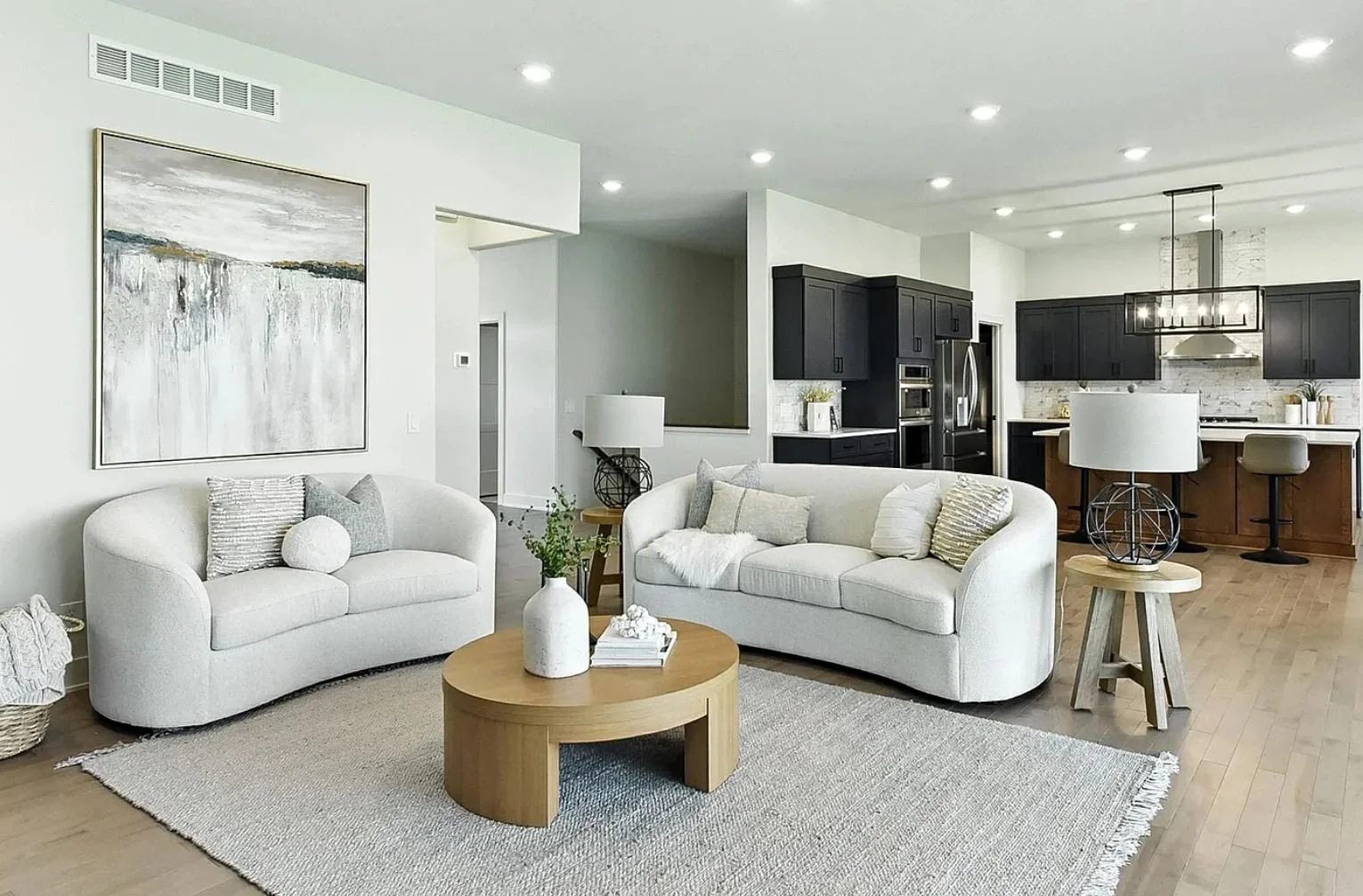 Modern living room with two white sofas, wooden coffee table, wall art, and an open kitchen with black cabinets and a marble backsplash.