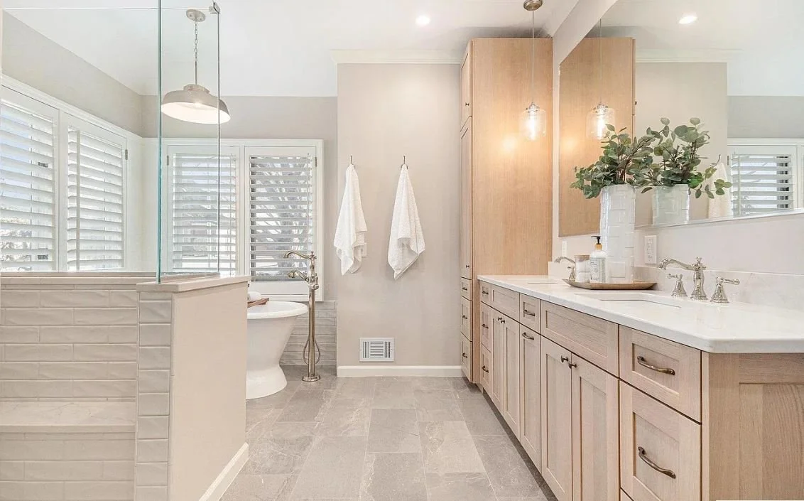 Bathroom with a light wood vanity, white countertop, two white towels hanging on the wall, a bathtub with a free-standing faucet, and white plantation shutters on the windows.